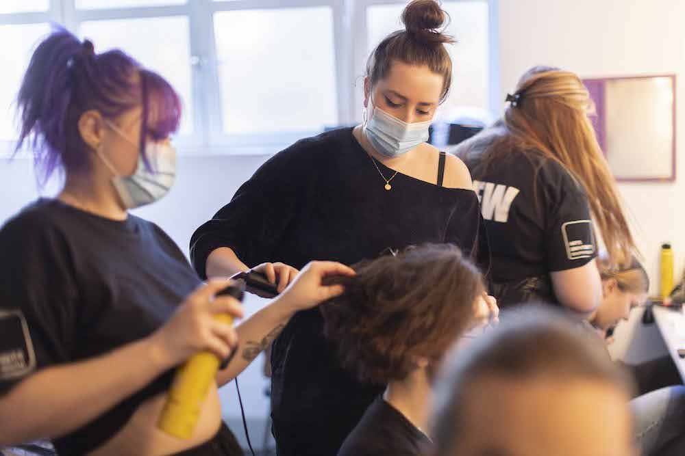 Two make-up artists wearing face masks work on an actor's hair.