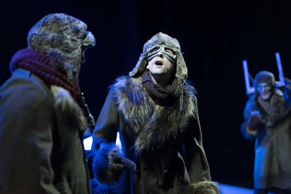 A performer in a fur-collared coat and hat and goggles gazes up open-mouthed.
