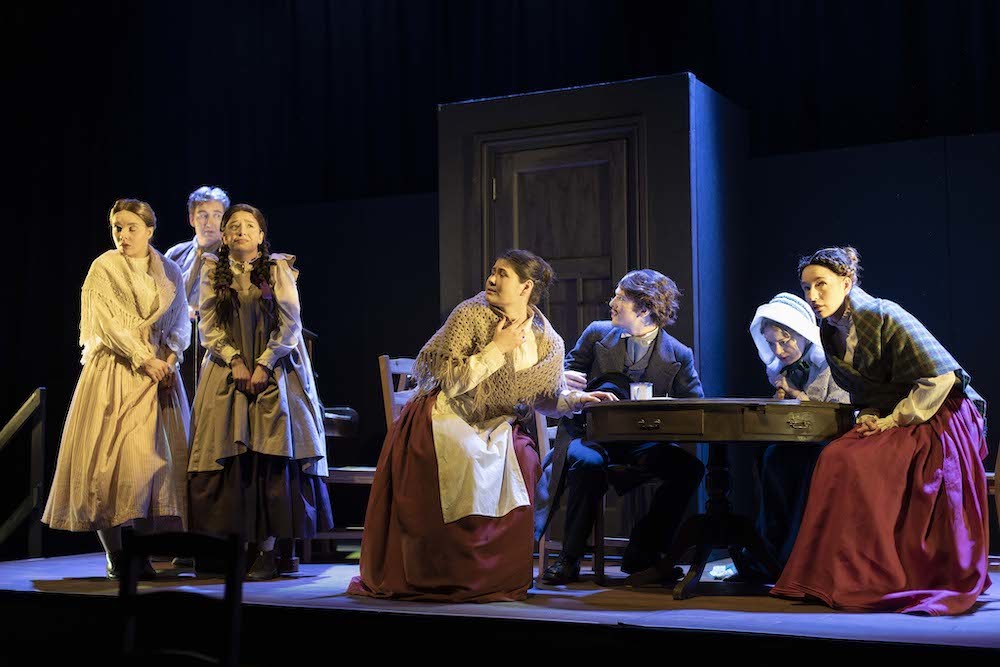 A group of actors in period costume in the middle of a performance. Four of them are seated around a table with a door behind them.