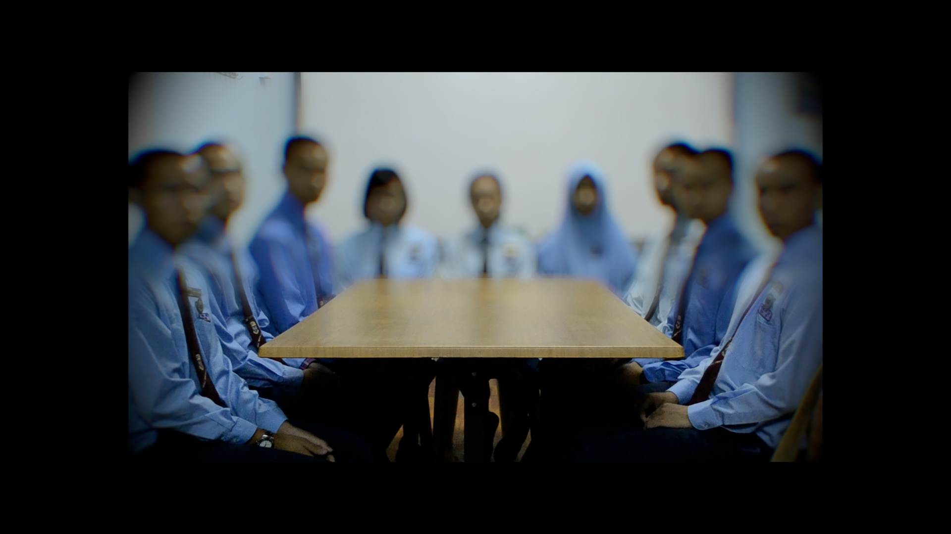 A group of office workers in blue shirts sit around a wooden conference table in a meeting room with a blurred white wall in the background.
