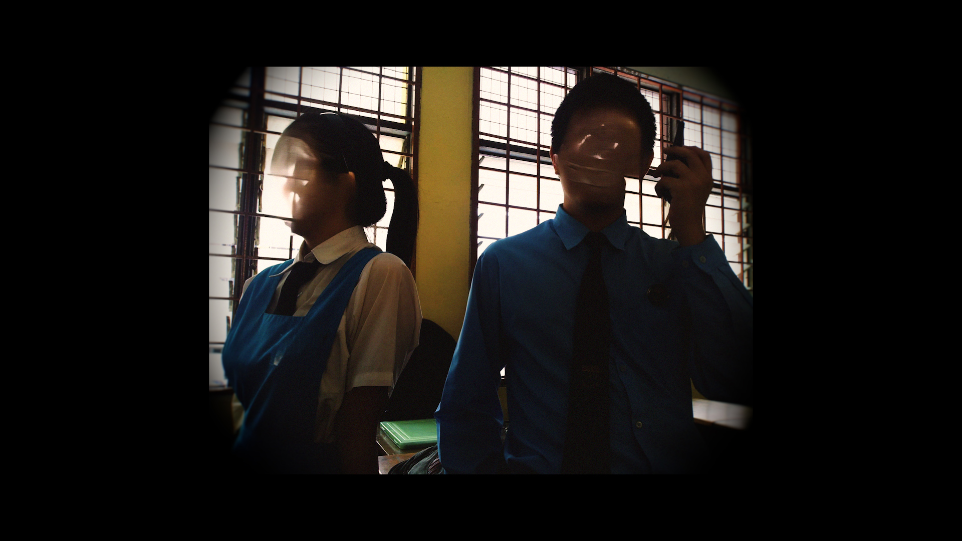 A woman in a blue apron and a man in a blue shirt stand indoors by barred windows, with bright daylight behind them.