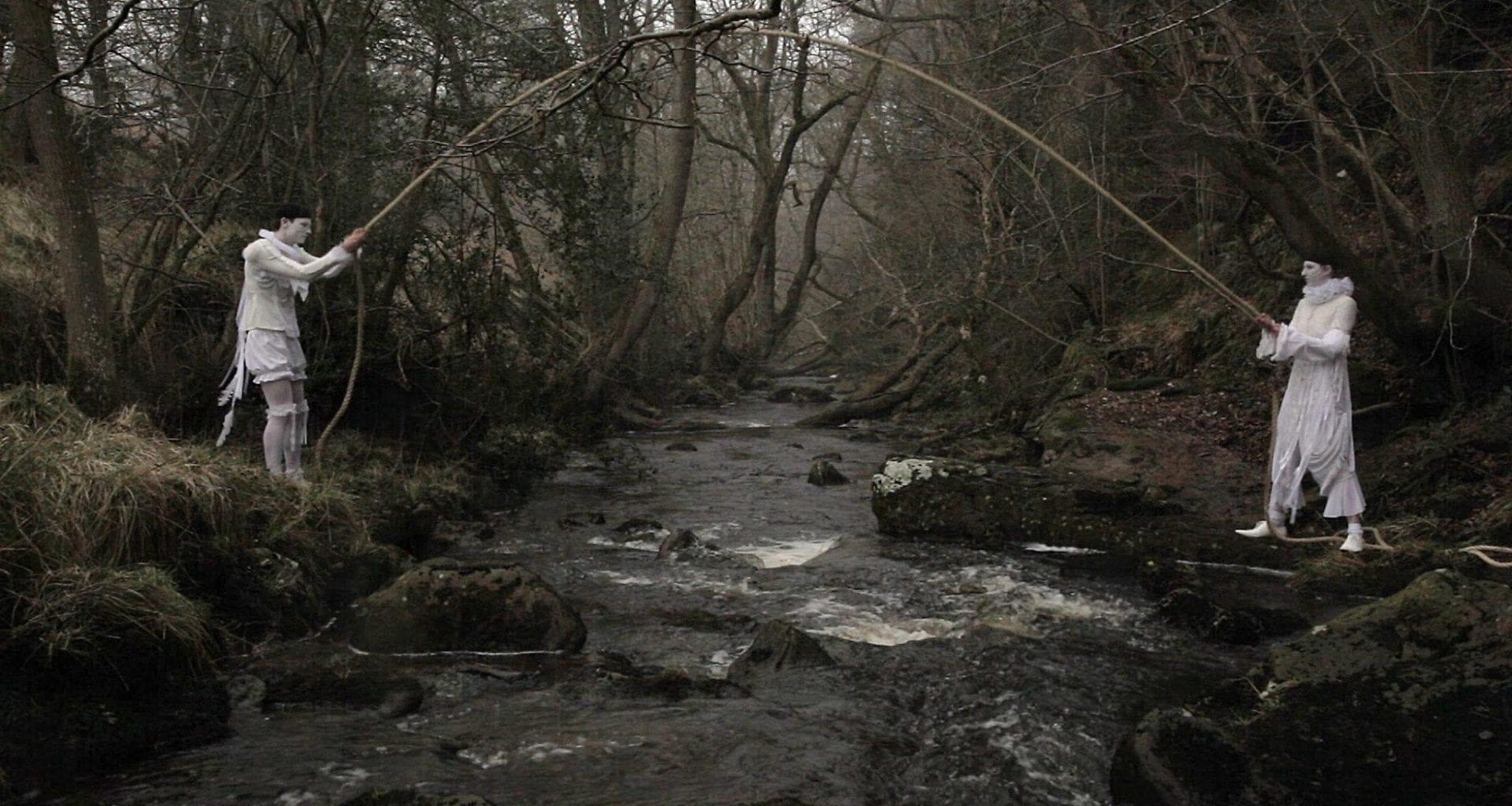 Two people in costume standing either side of a river in a woodland and each holding one end of a long pole.