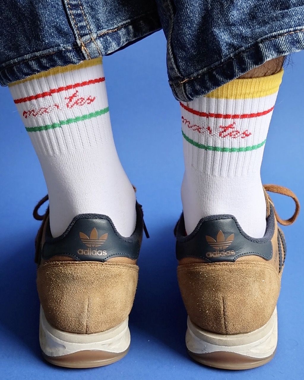 A person wearing tan Adidas trainers with white socks and denim jeans, shown from behind against a blue background.