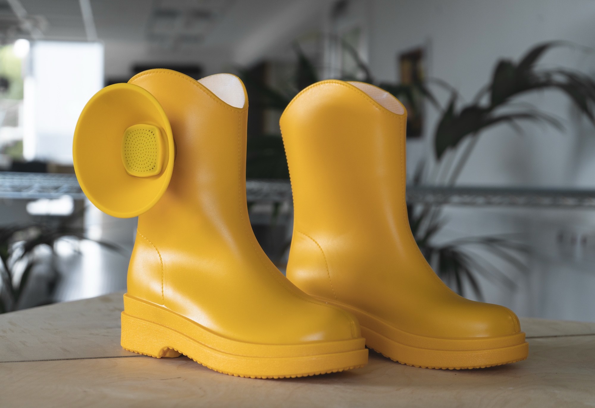 A pair of bright yellow rubber boots with rounded toes, placed on a wooden surface with a blurred indoor background.