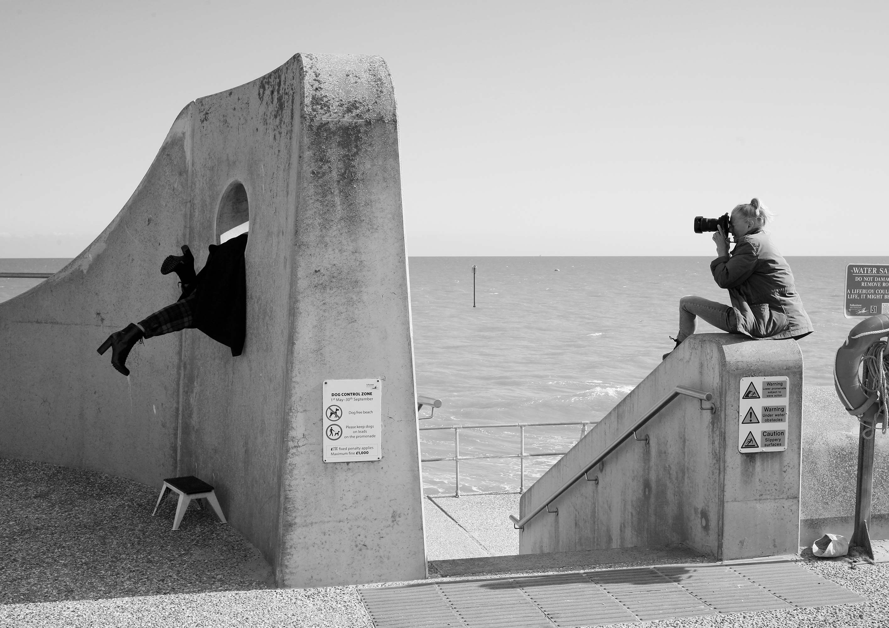 Black and white photograph of W Carrig taking a photo of another person whose legs are sticking out of a hole in a wall by a beach.
