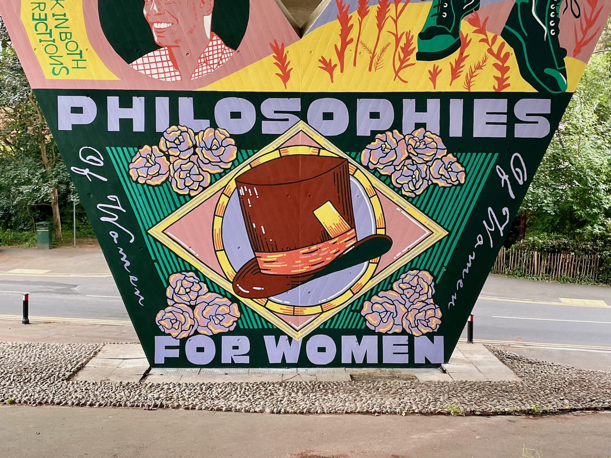 Part of a mural on a flyover pillar wall, primarily green quadrangle with a pink diamond and a brown top hat in the centre. Text appears along the top and bottom reading "Philosophies for women", and "Of women" along either side.