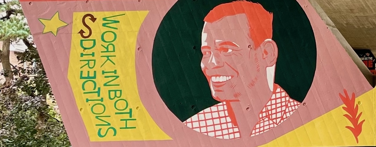 Part of a mural on a flyover pillar, depicting a smiling man in a black circle. A yellow box to the left contains text reading "Works in both directions" with some letters inverted.