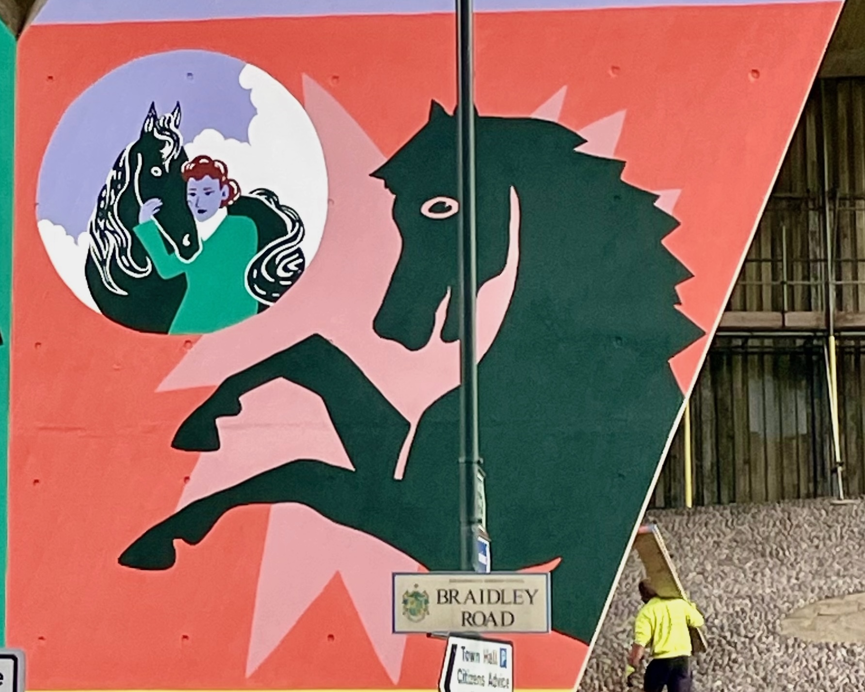 Part of a mural on a flyover pillar wall. Part of a horse in silhouette against a red and pink background. A smaller illustration of a woman with a black horse is show in a purple and white circle in the left corner.