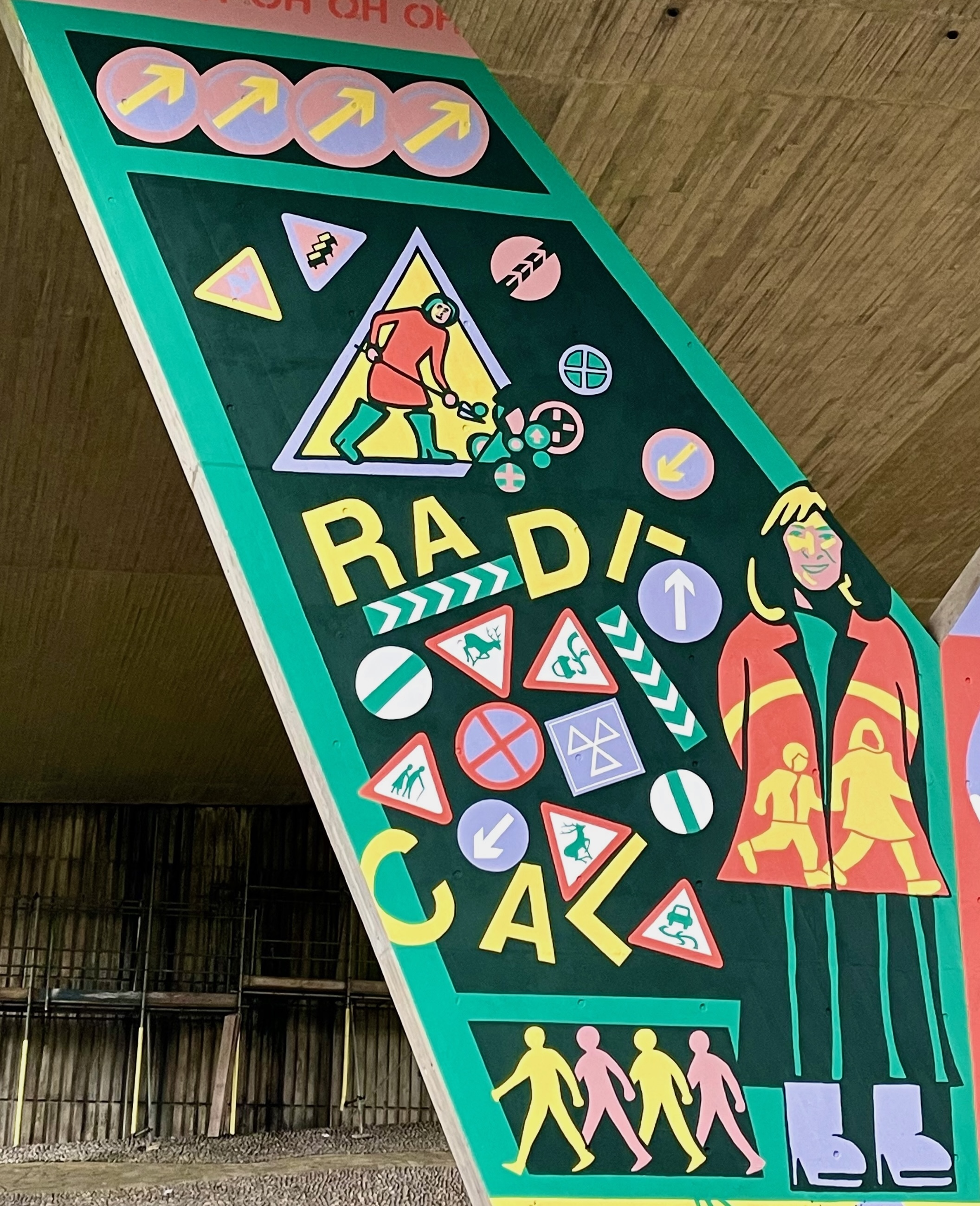 Part of a mural on a flyover pillar wall; an assortment of road signs and the word "RADICAL". An illustration of a woman wearing a hi-vis jacket is shown on the right.