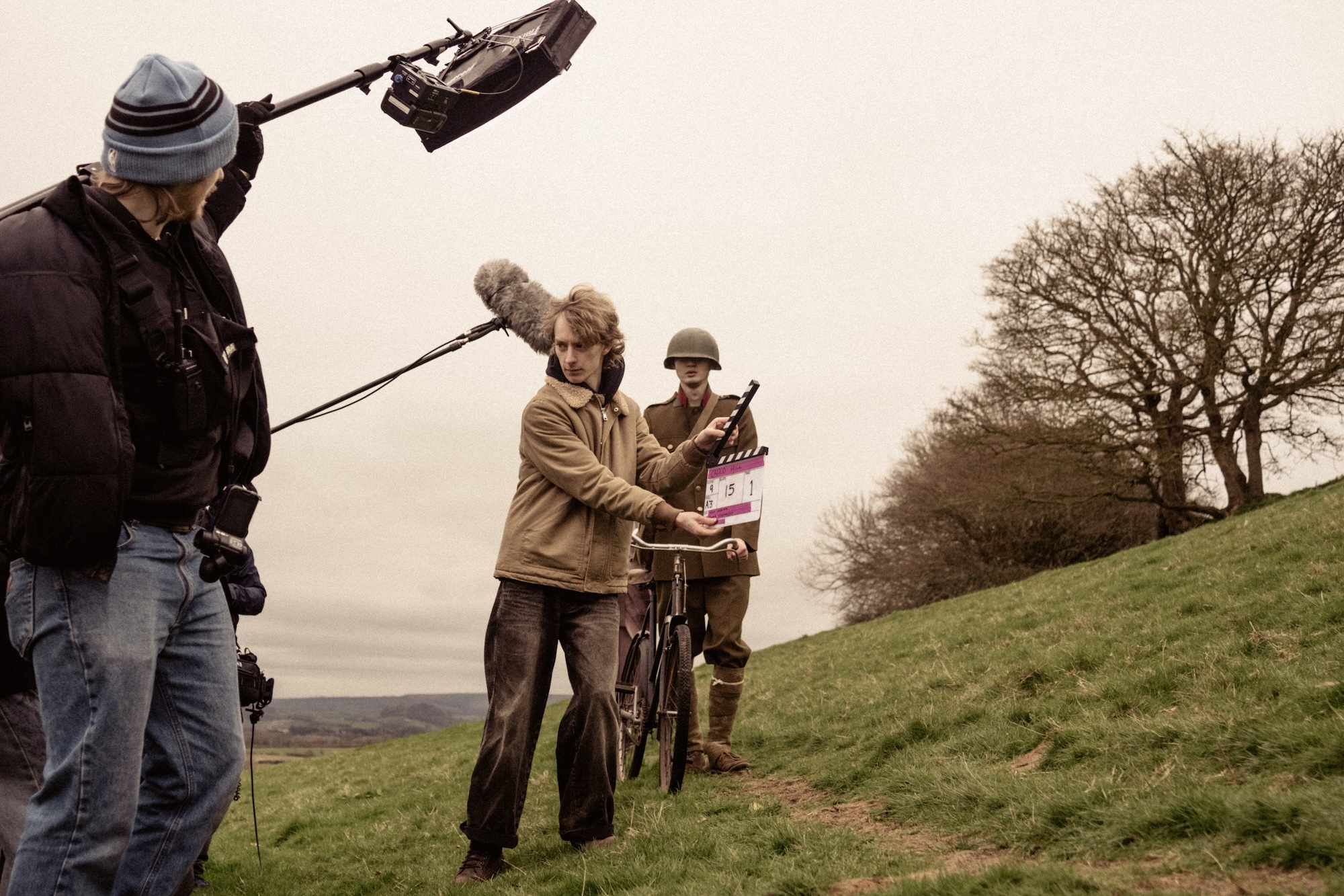 A film crew is on a grassy hillside, with one member holding a clapperboard and bicycle, while another operates a boom microphone. A man dressed in a military uniform stands beside them, and trees are visible in the background.