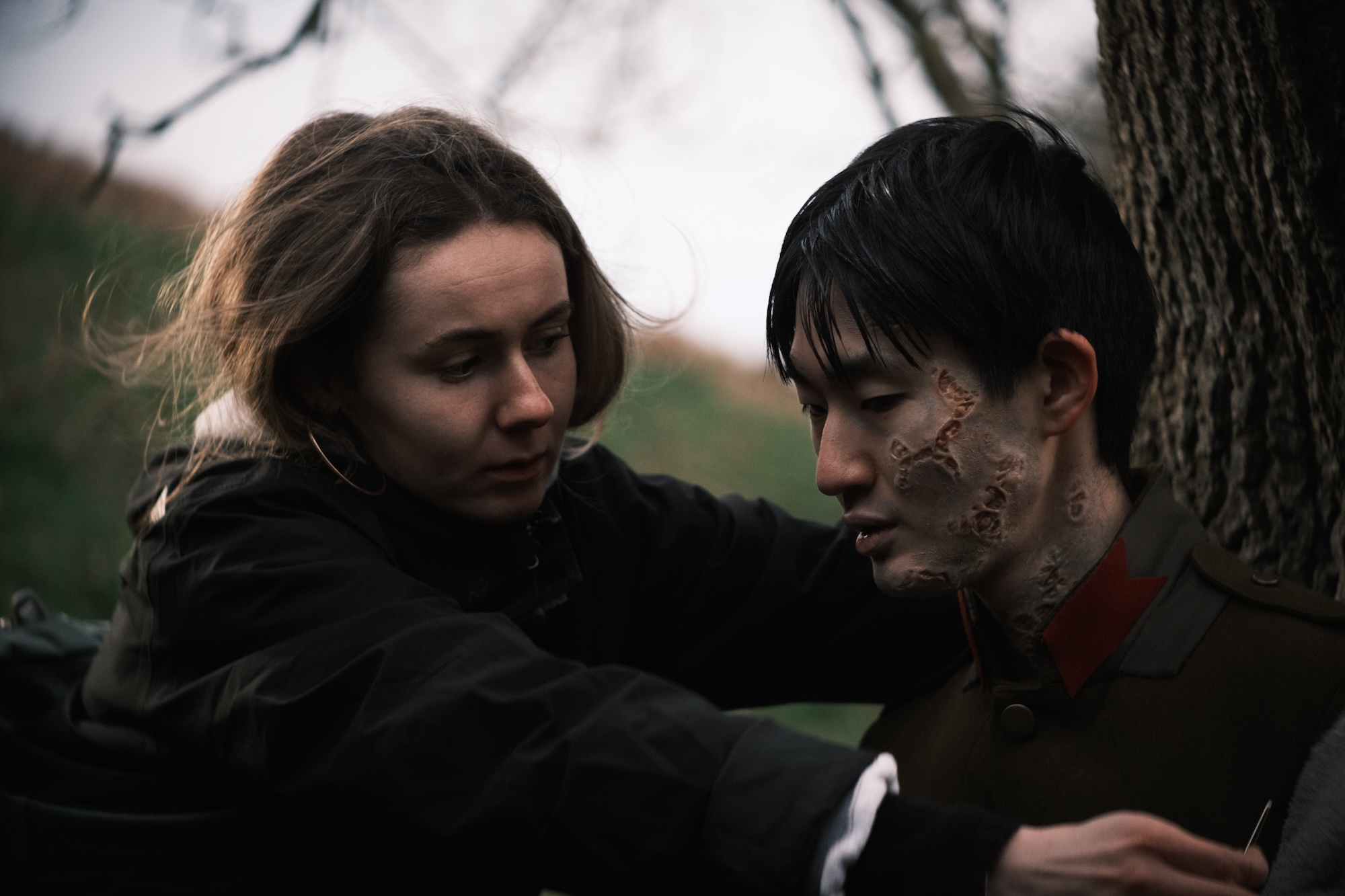 A woman with long brown hair gently touches the shoulder of a man with short black hair and visible scars on his face. They are outdoors, with a tree trunk partially visible in the background and a grassy area.