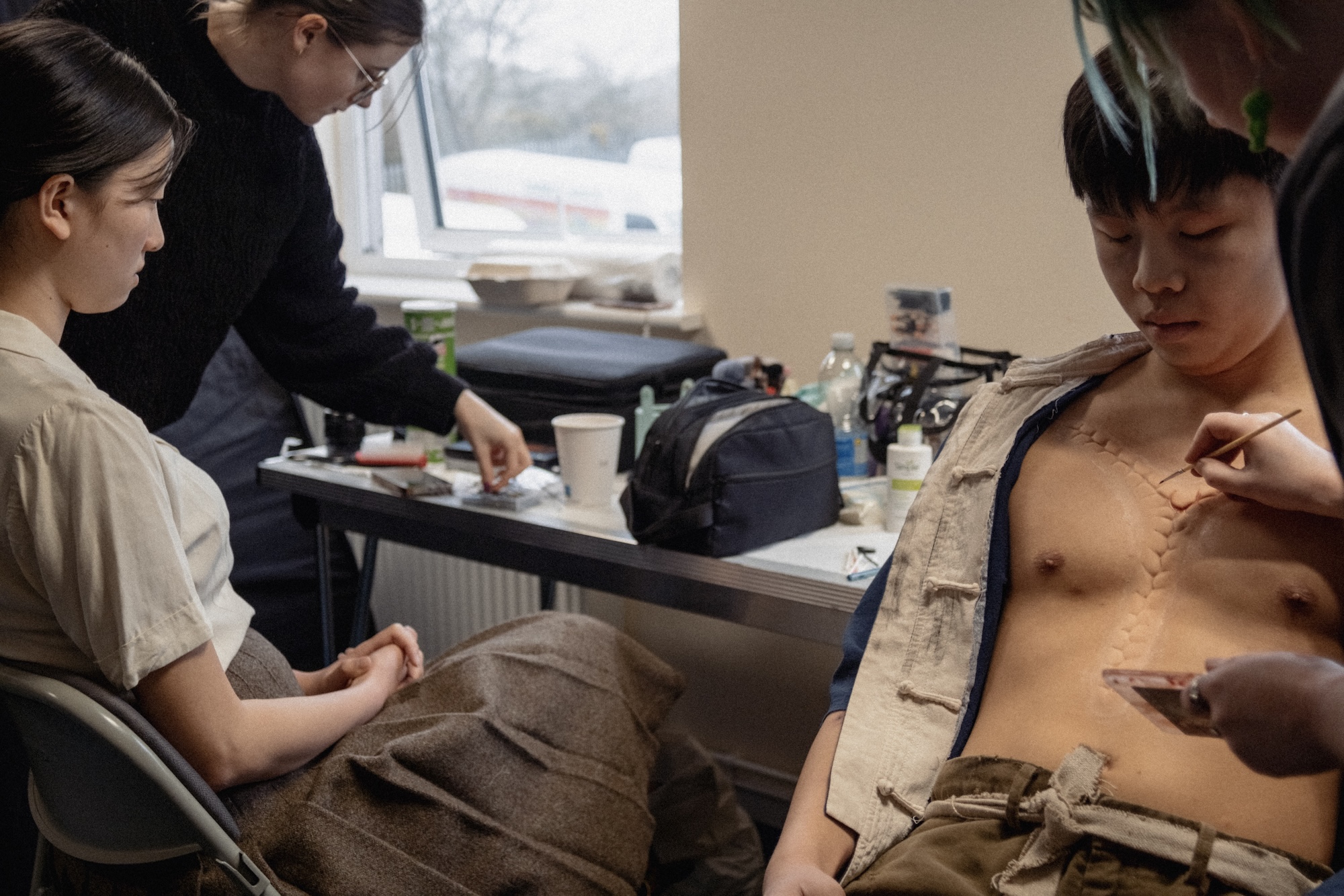A woman sits on a chair, wearing a beige outfit, while a man with short dark hair sits next to her. He has a make-up artist applying effects on his bare chest, which has a detailed artificial scar. A table cluttered with makeup tools is in the background.