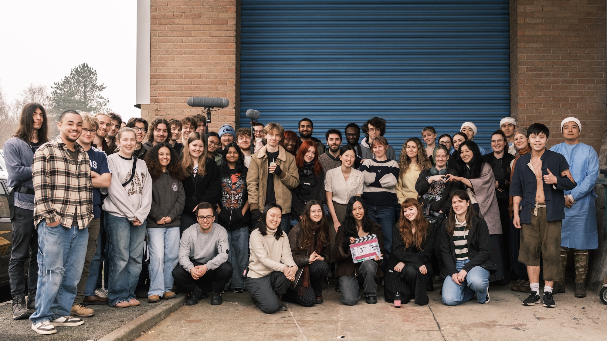 A diverse group of around 40 people stand outdoors in front of a blue shuttered garage door. They are dressed casually and a few hold film equipment, including a clapperboard. The setting appears to be a filming location.