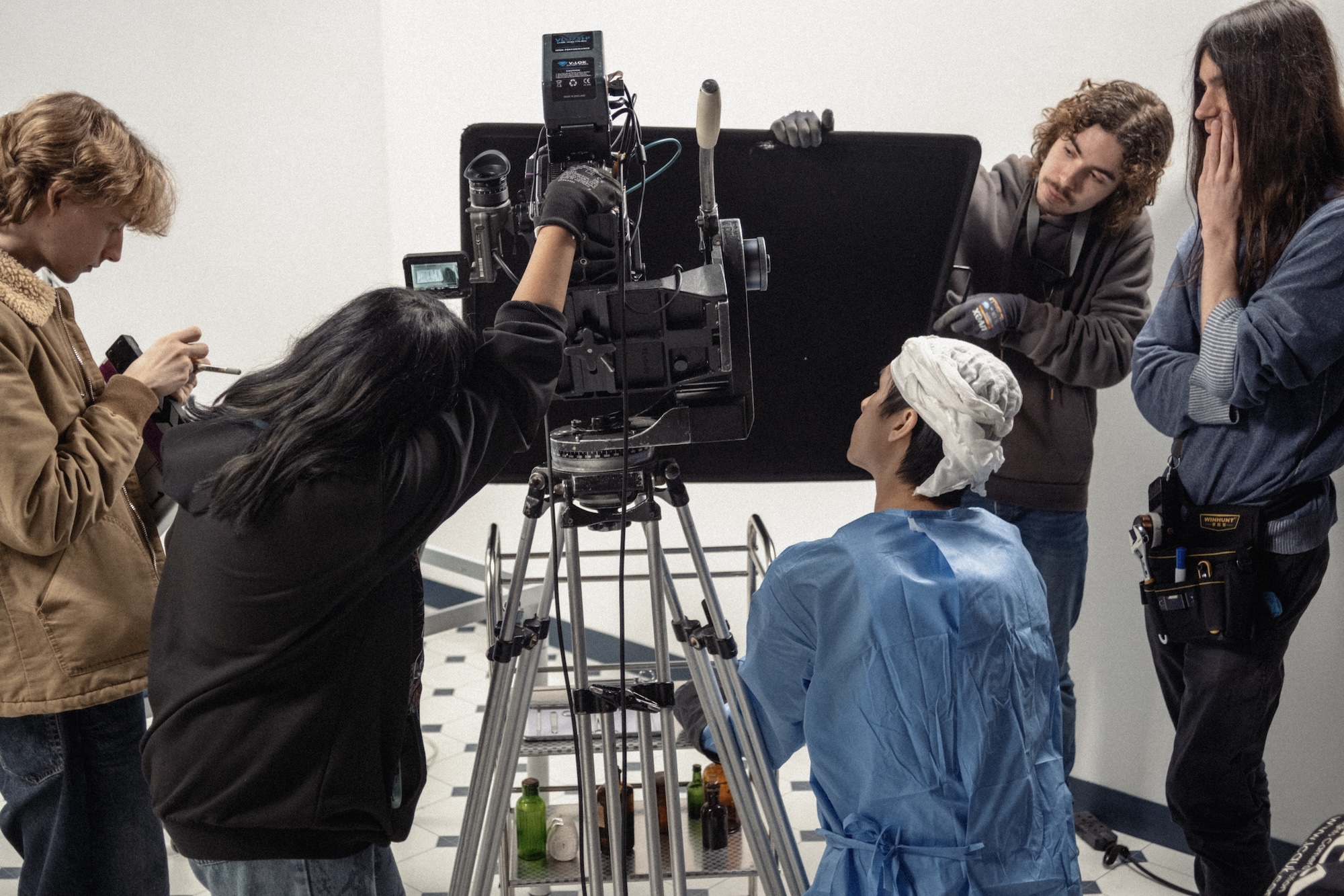 A crew of five people is gathered around a large film camera on a tripod. One individual is adjusting the camera, while others are observing and preparing equipment, with a focus on a man in medical scrubs and a surgical cap.