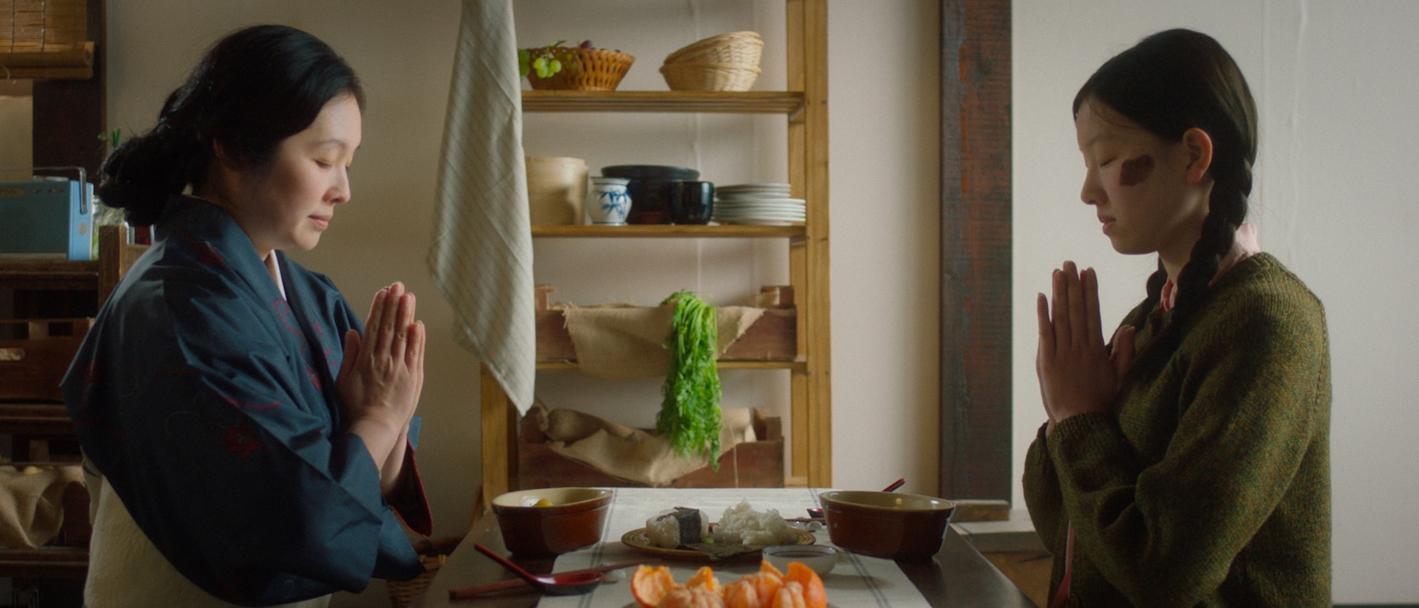A woman in a traditional robe and a girl in a green sweater are seated at a table with their hands clasped in prayer. In front of them are bowls of rice, a plate of sliced fruit, and a backdrop of wooden shelves with kitchen items.