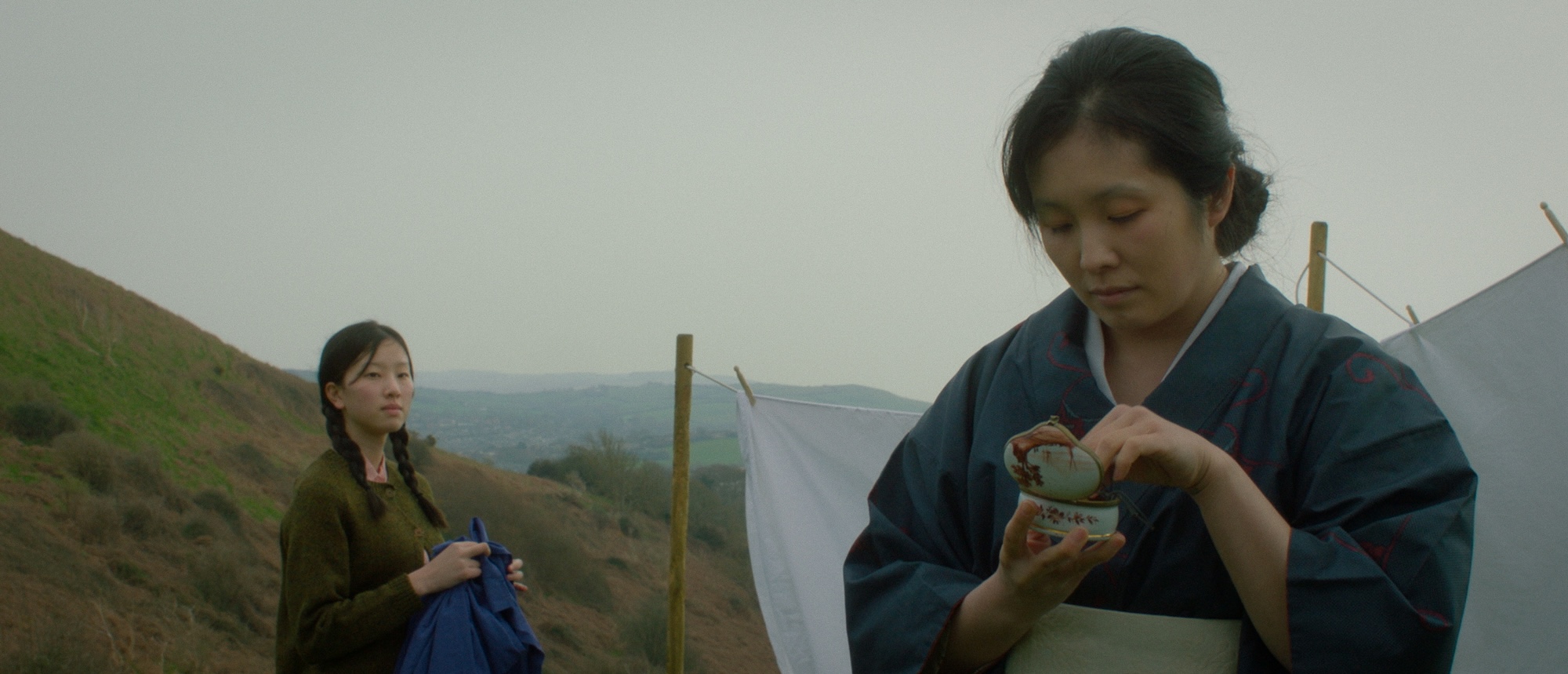 Two women in a landscape setting. One woman, dressed in a traditional kimono, is holding a decorative container while focusing on its contents. The other woman, wearing a dark jacket, stands in the background, holding a blue garment, with hills visible behind them.
