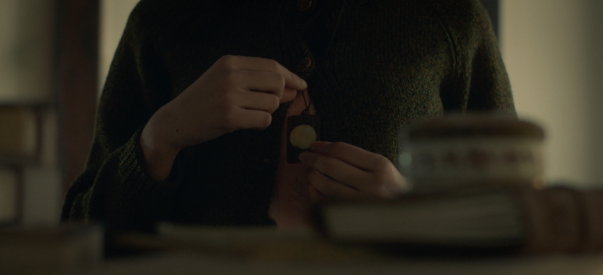 A hand holding a small pendant is visible, with the person wearing a dark green knitted cardigan. The background includes blurred books and a container on a wooden surface.