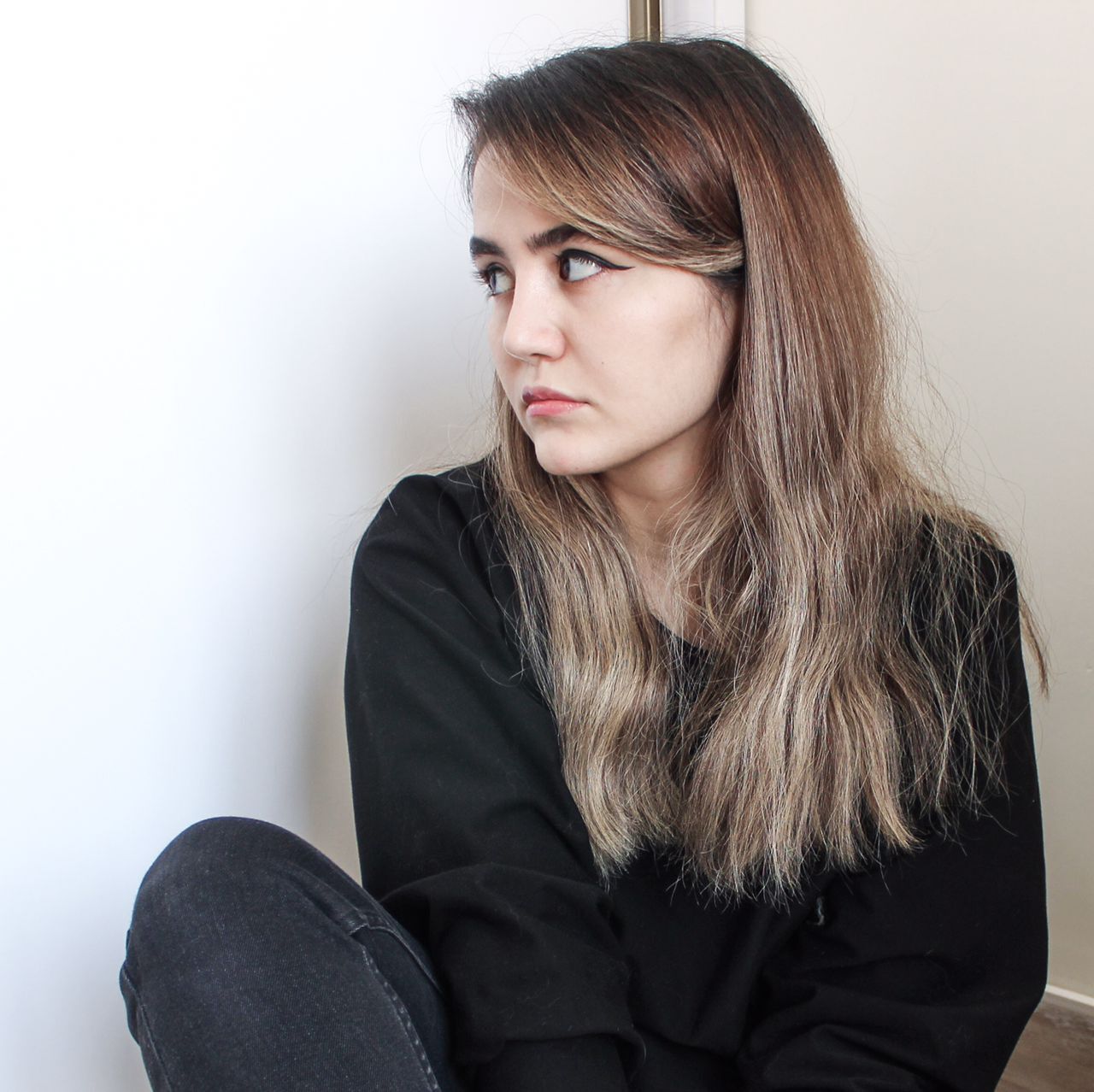 Woman with long brown hair and side fringe wearing black jumper, looking to the right whilst sitting against white wall.