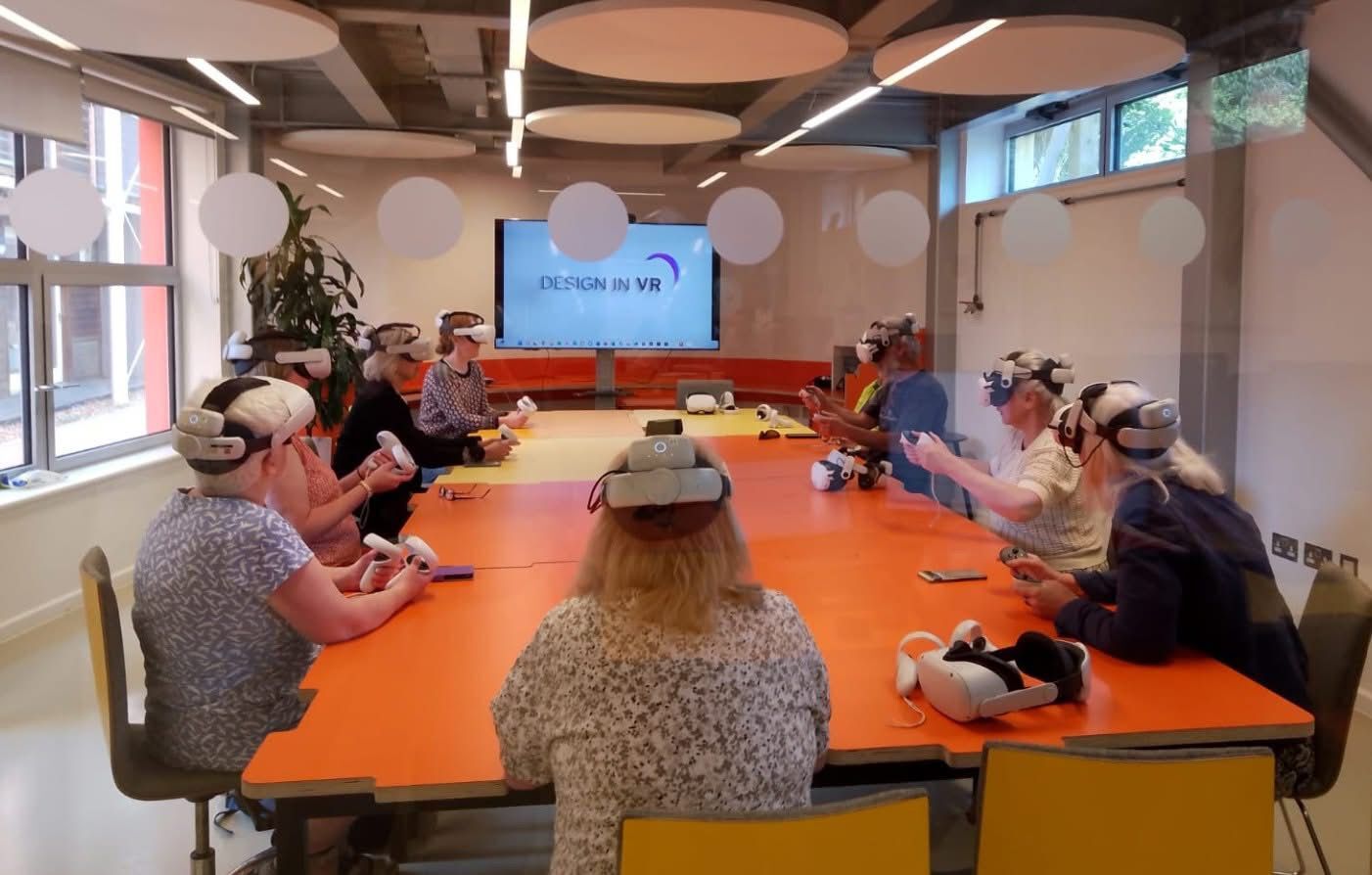A group of Bournemouth, Christchurch and Poole councillors sitting around a large table in the board room at the Innovation Studio, Arts University Bournemouth, wearing VR headsets. A screen at the far end of the room reads "Design in VR".
