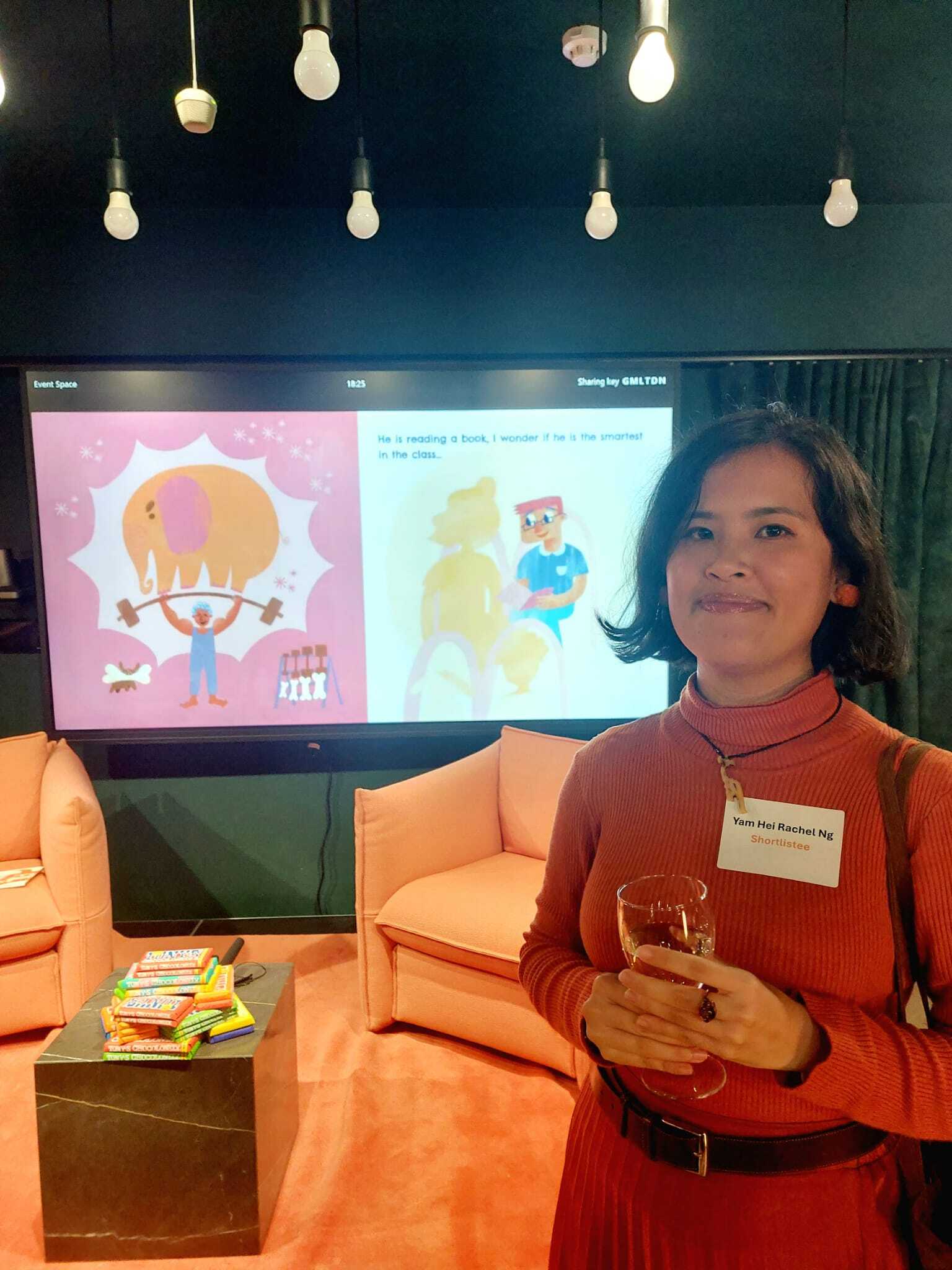 R Ng at the 2025 FAB Awards, standing in front of an orange armchair holding glass, with large screen showing colourful children's book illustrations behind her.
