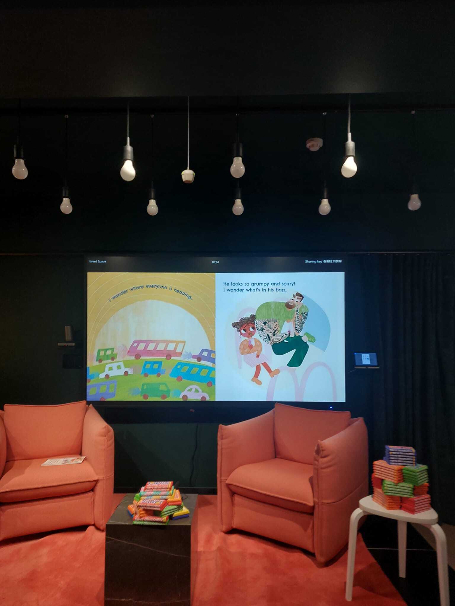Dark room with hanging bulbs, two orange armchairs facing large screen displaying colourful children's book illustrations.