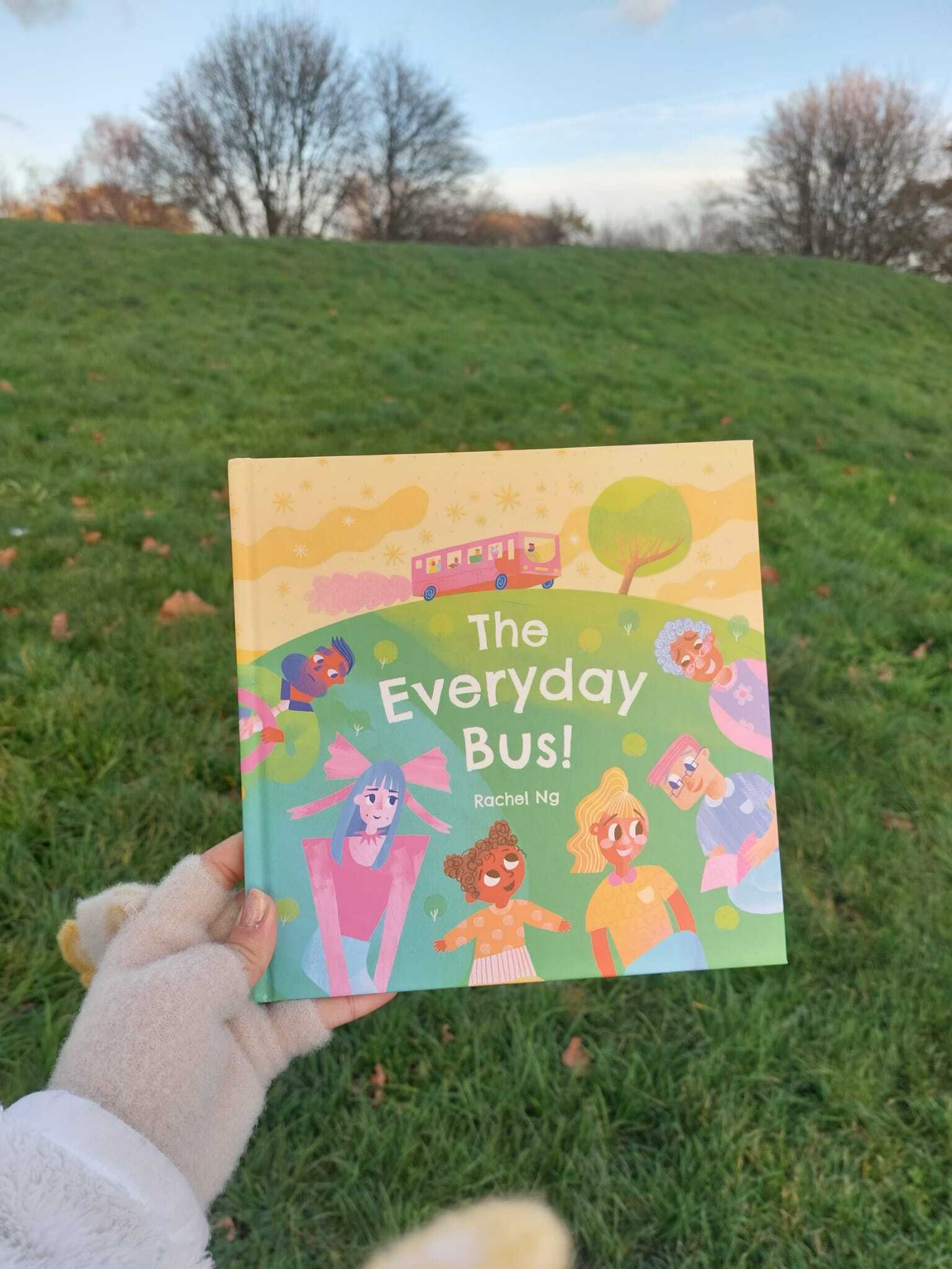 Hand holding "The Everyday Bus!" children's book with colourful illustrated cover featuring cartoon characters, against green grass field.