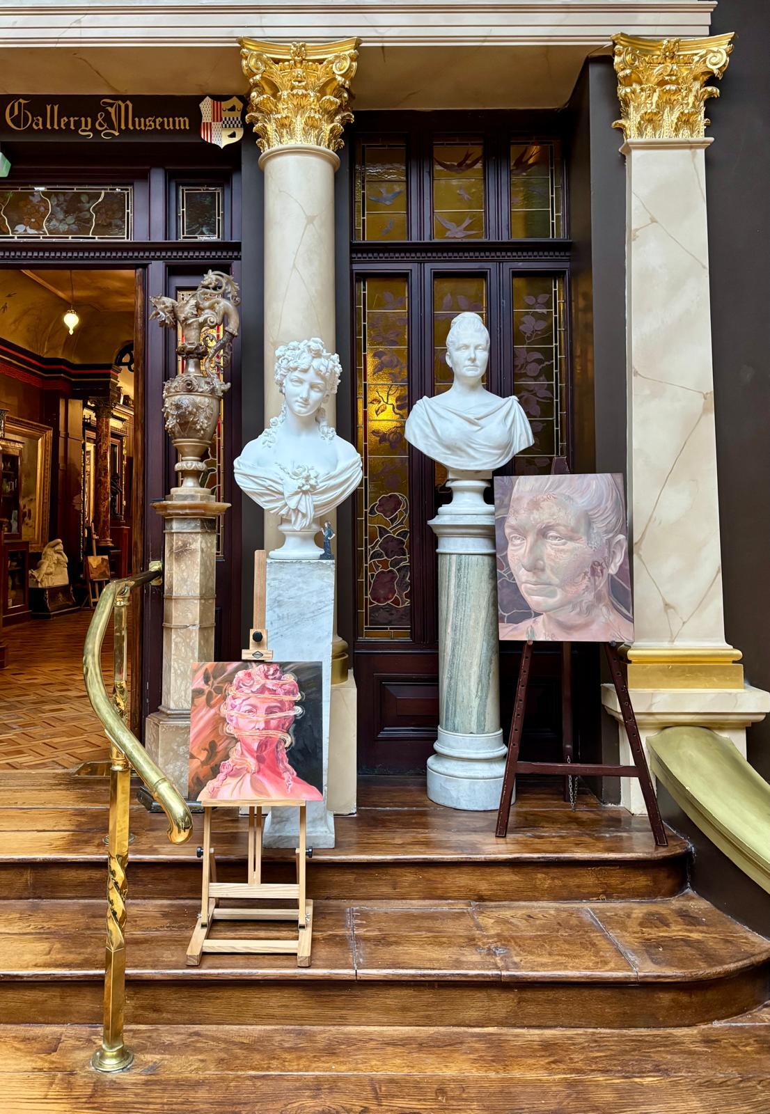 Gallery interior with white marble columns, gold Corinthian capitals, dark wood panelling, white classical busts, and pink portrait paintings on easels.