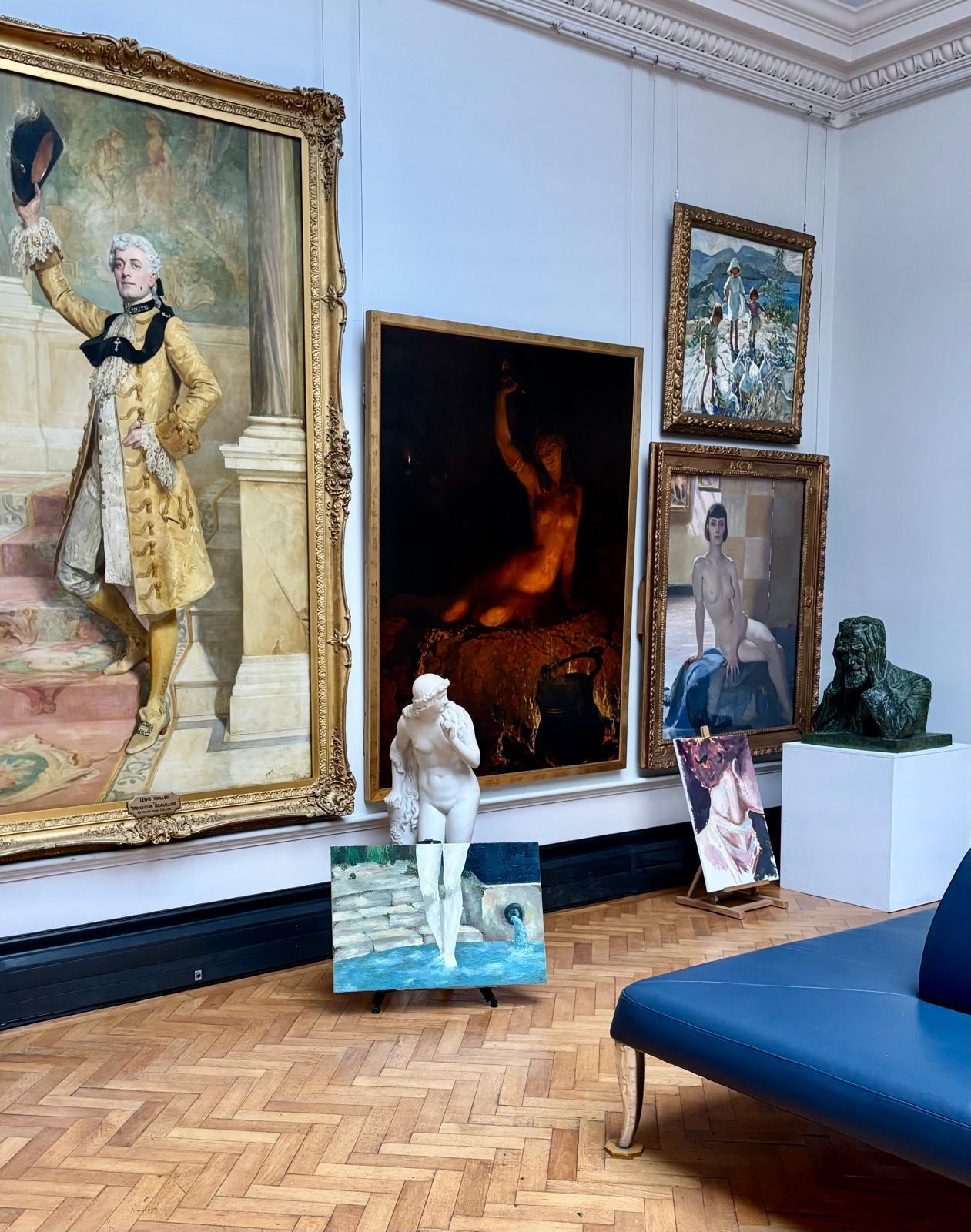 Gallery room with ornate paintings on white walls, parquet flooring, blue bench, white sculpture on pedestal, decorative ceiling moulding.