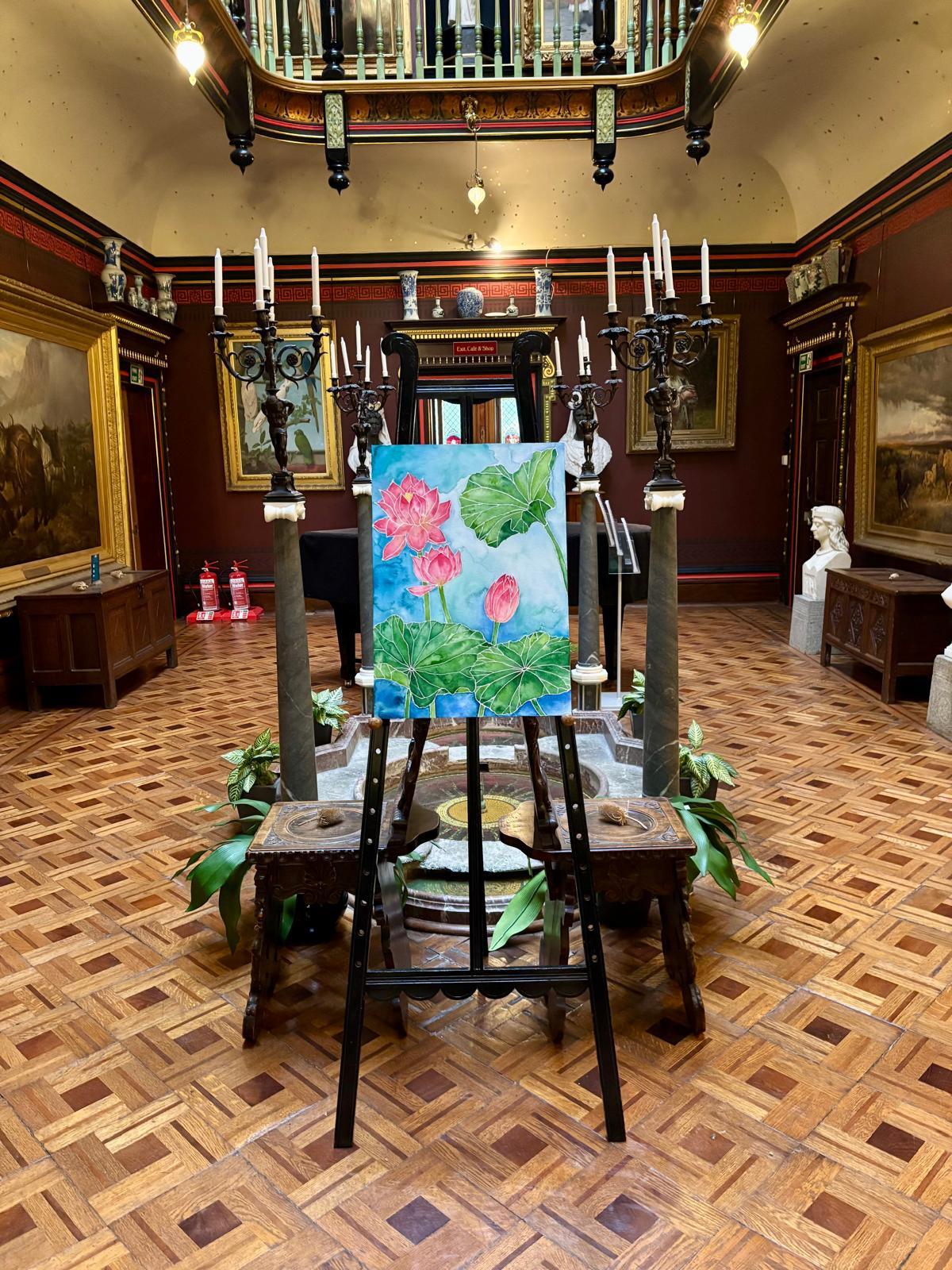 Black wooden easel holds canvas with pink flowers and green foliage against blue background in ornate gallery with red walls, parquet flooring.