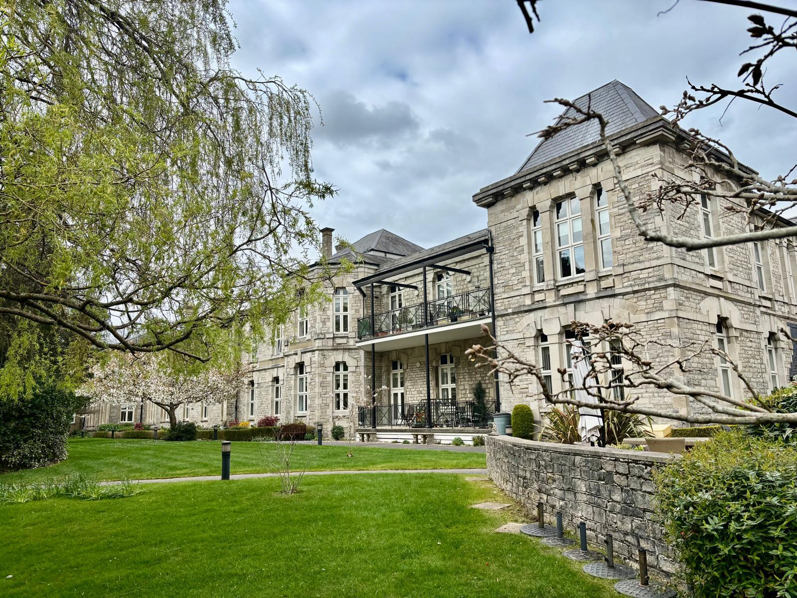 Large stone mansion with grey slate roof and iron balconies, surrounded by green lawn and mature trees under cloudy sky