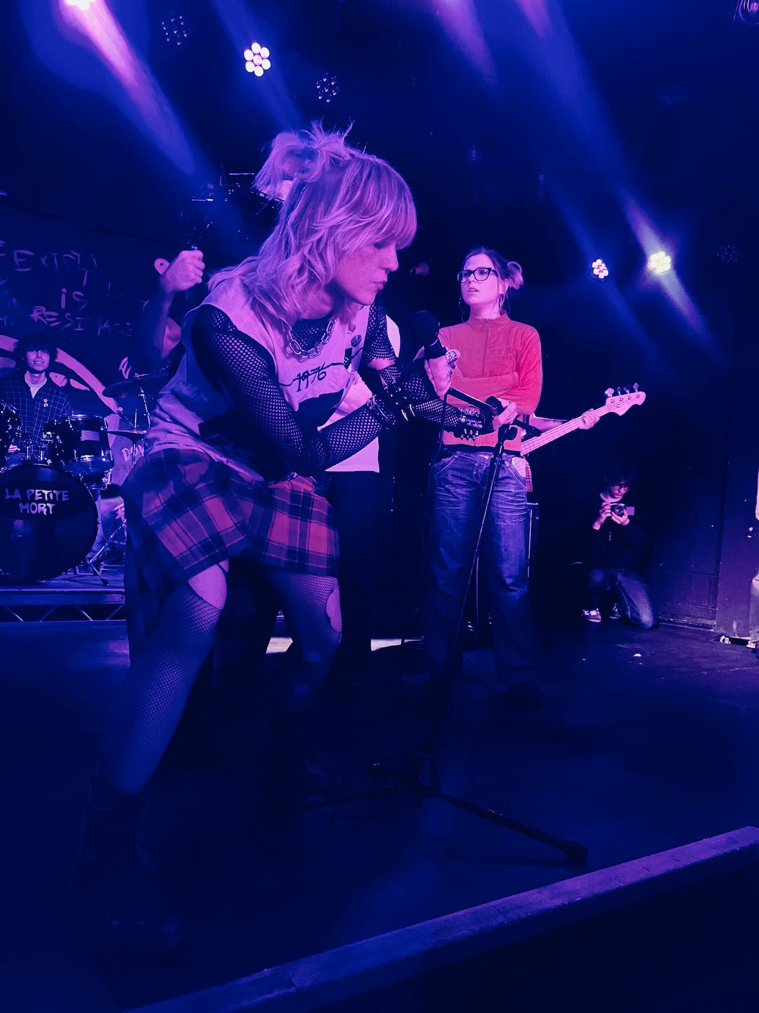 A female performer in a punk-style outfit, including a short plaid skirt and fingerless gloves, leans into a microphone on stage. Behind her, a band plays with brightly lit instruments in a venue featuring dark walls and vibrant stage lights.