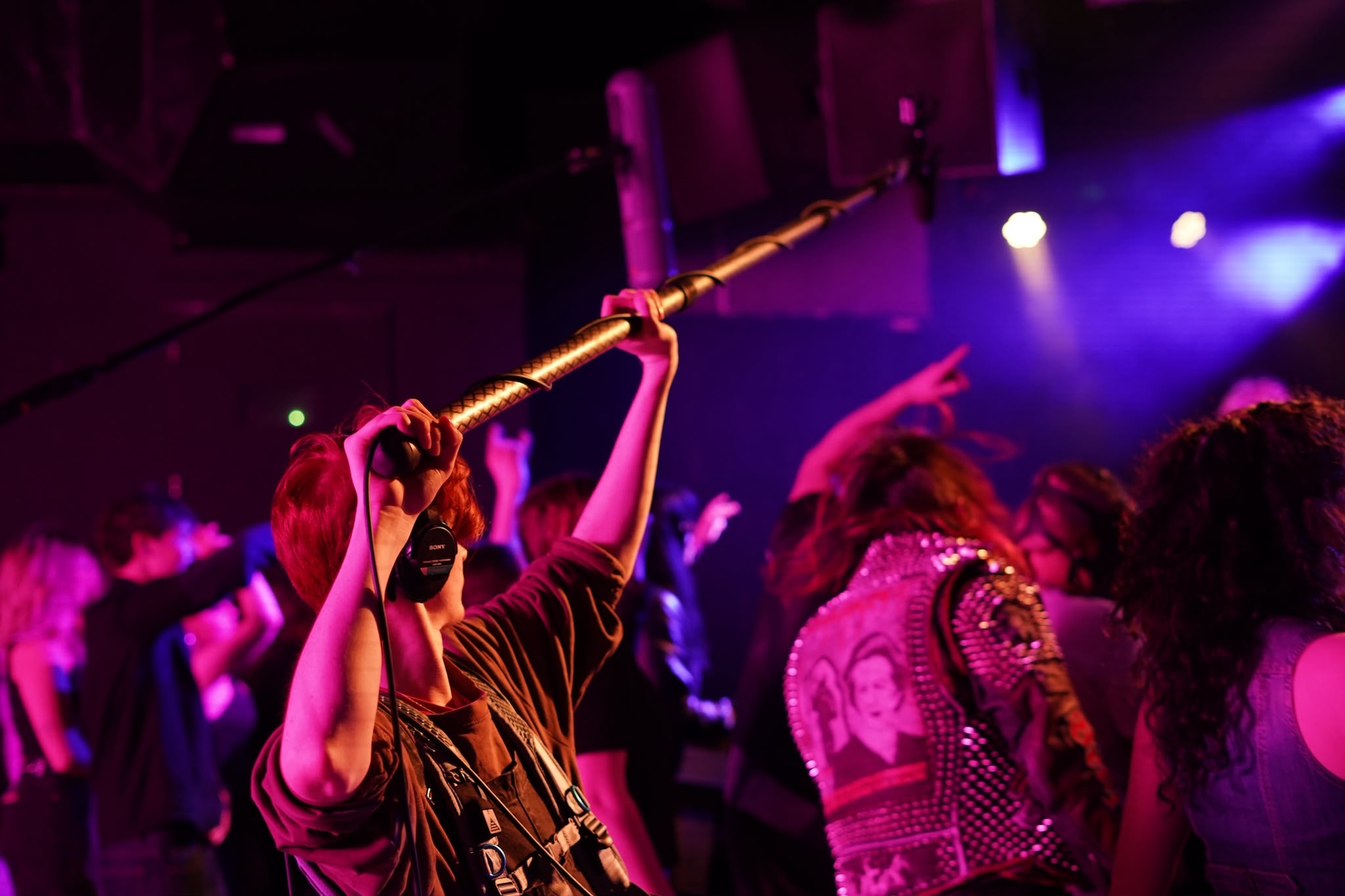 A crowd of people dancing in a dimly lit venue with purple and blue lighting. A person with short red hair holds a microphone on a long pole, while others are gesturing with their hands in an energetic atmosphere.