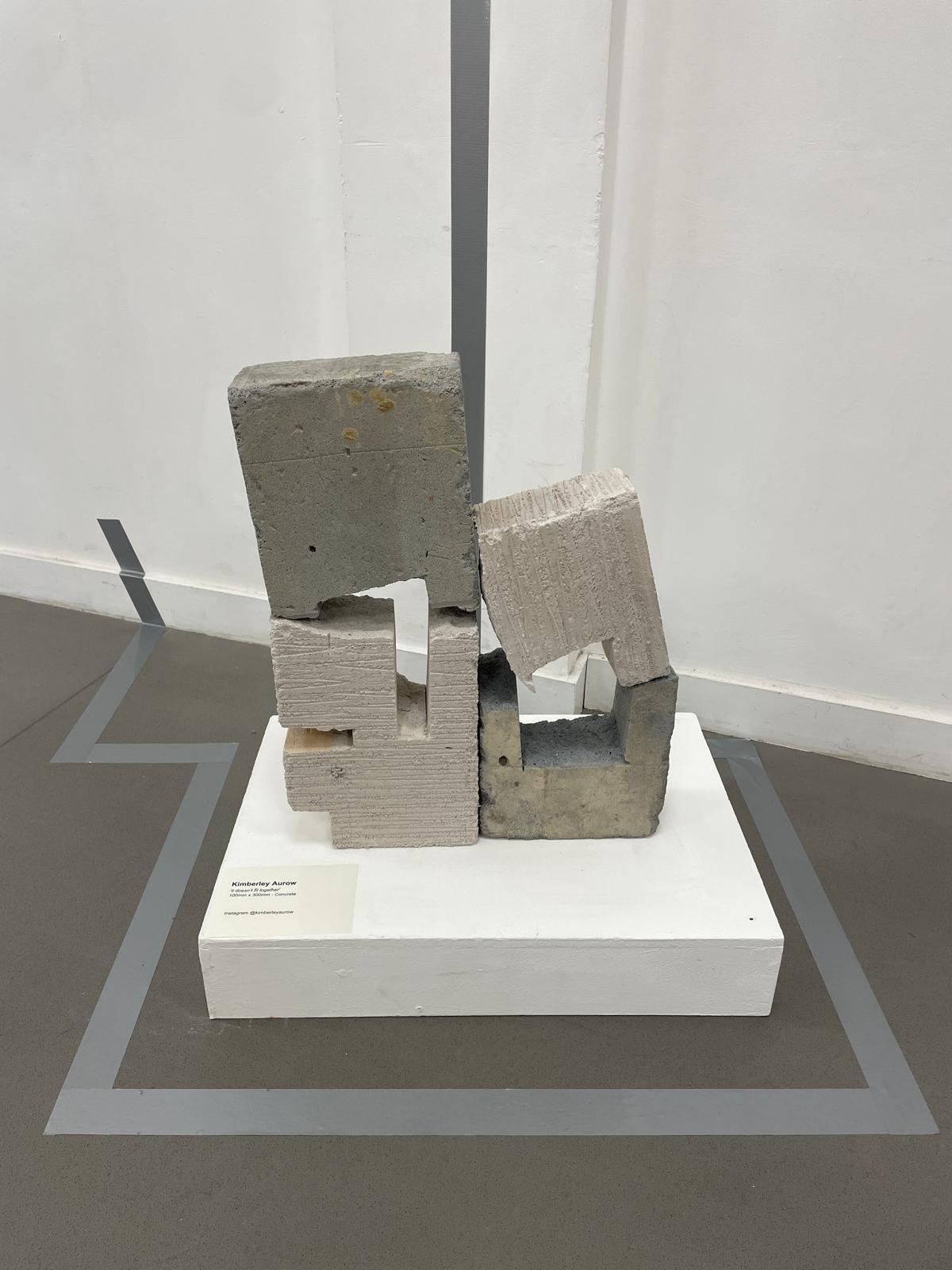 An abstract sculpture made of stacked grey concrete and textured blocks on a white plinth, displayed on a grey floor against a plain white wall.