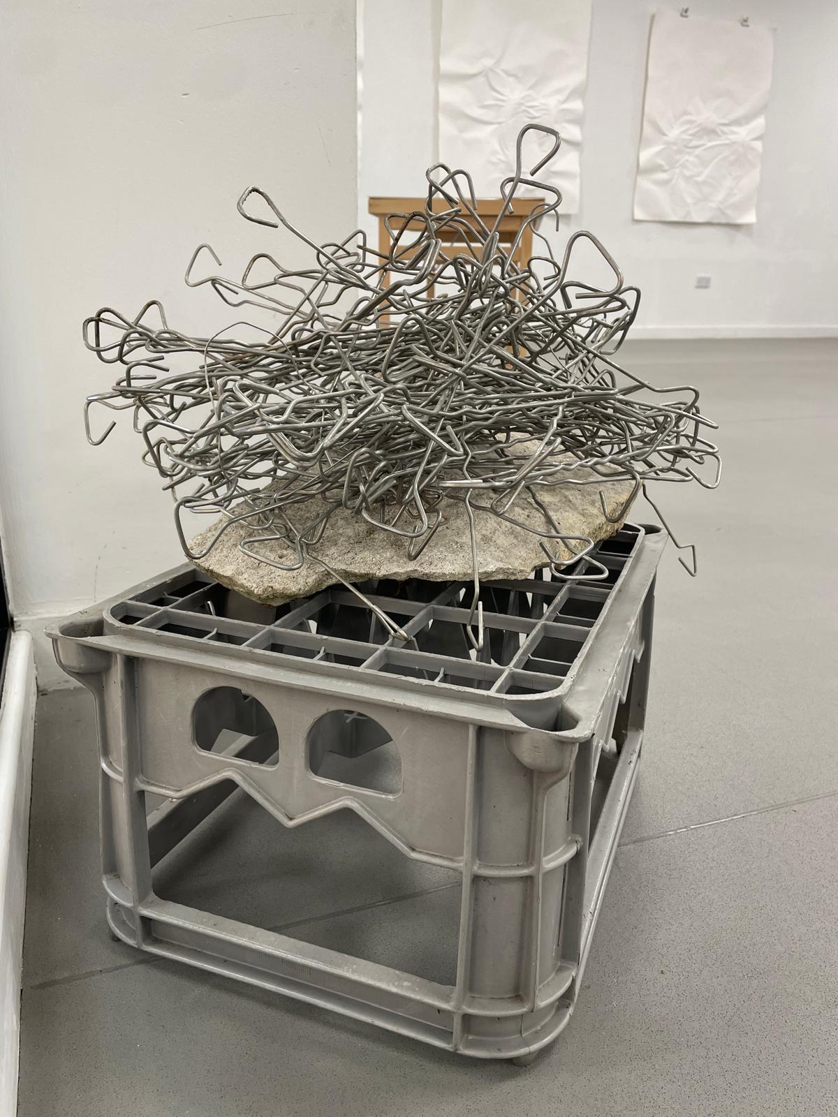 A pile of twisted metal wire rests on a rough stone slab balanced on a grey plastic crate in a white gallery space with framed wall pieces.