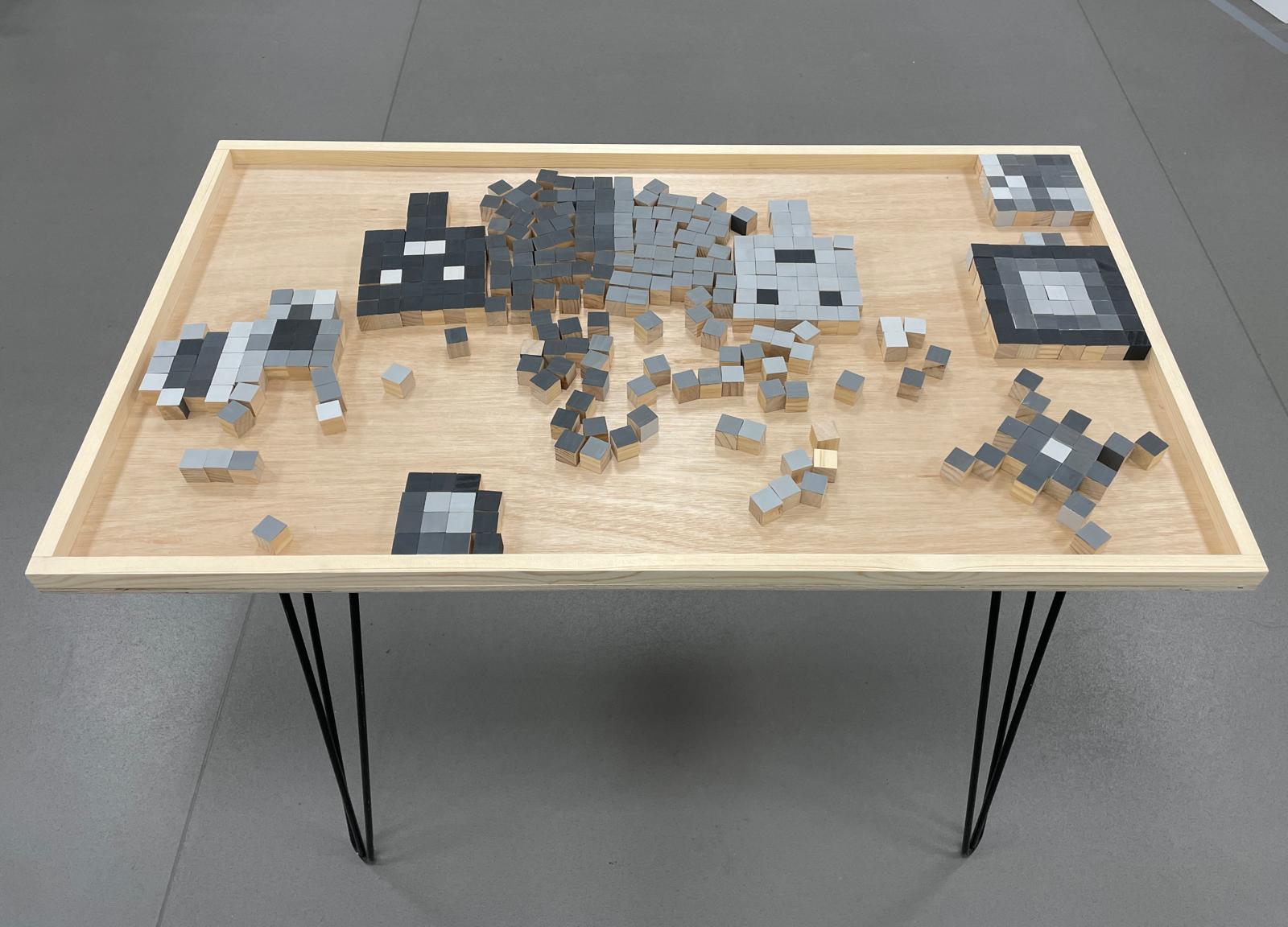 A pixelated mosaic artwork of a cat made from small grey, black and white tiles on a wooden table with thin black legs, set on a grey floor.