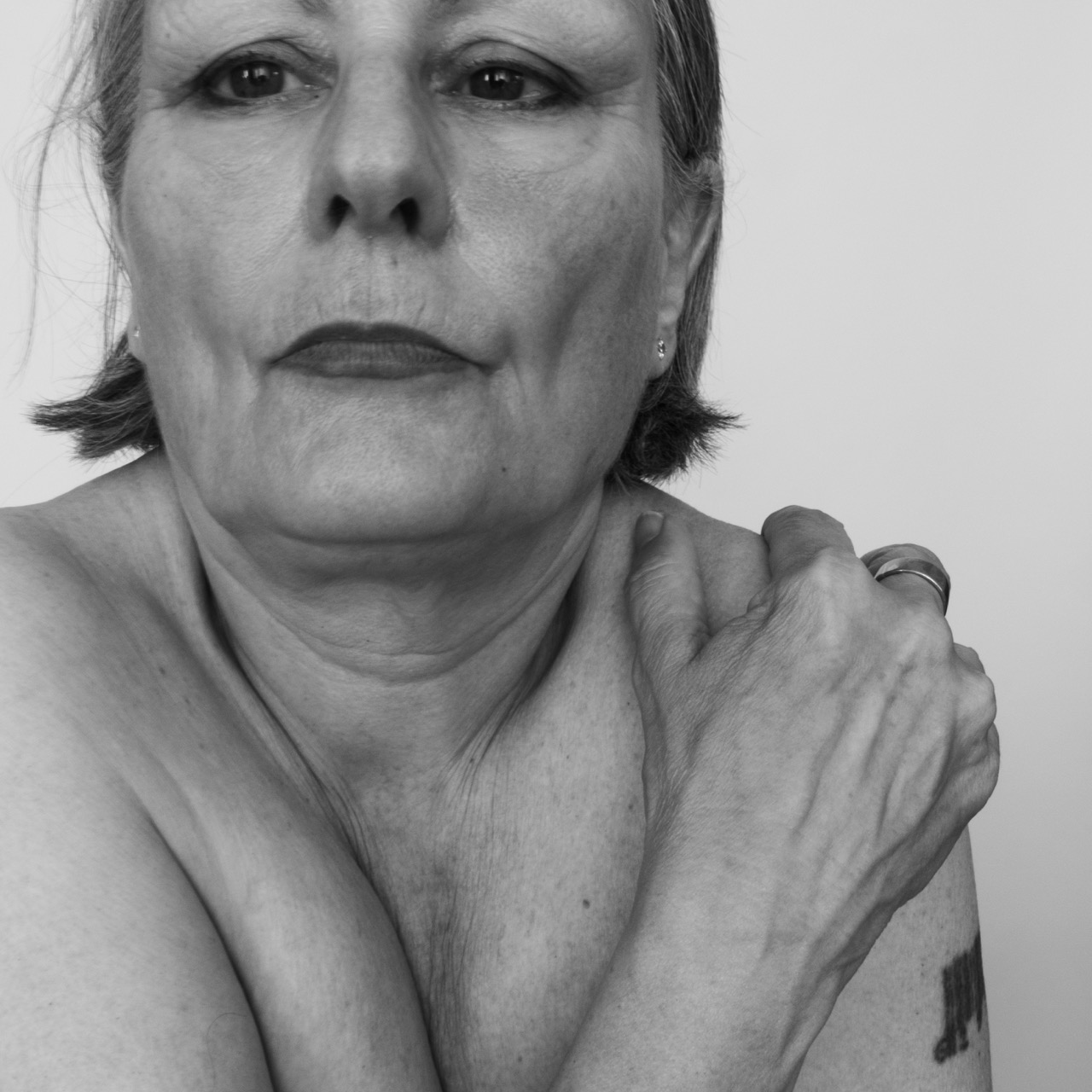 Black-and-white close-up of a woman with short hair holding her shoulder, with a ring visible on her hand against a plain background.