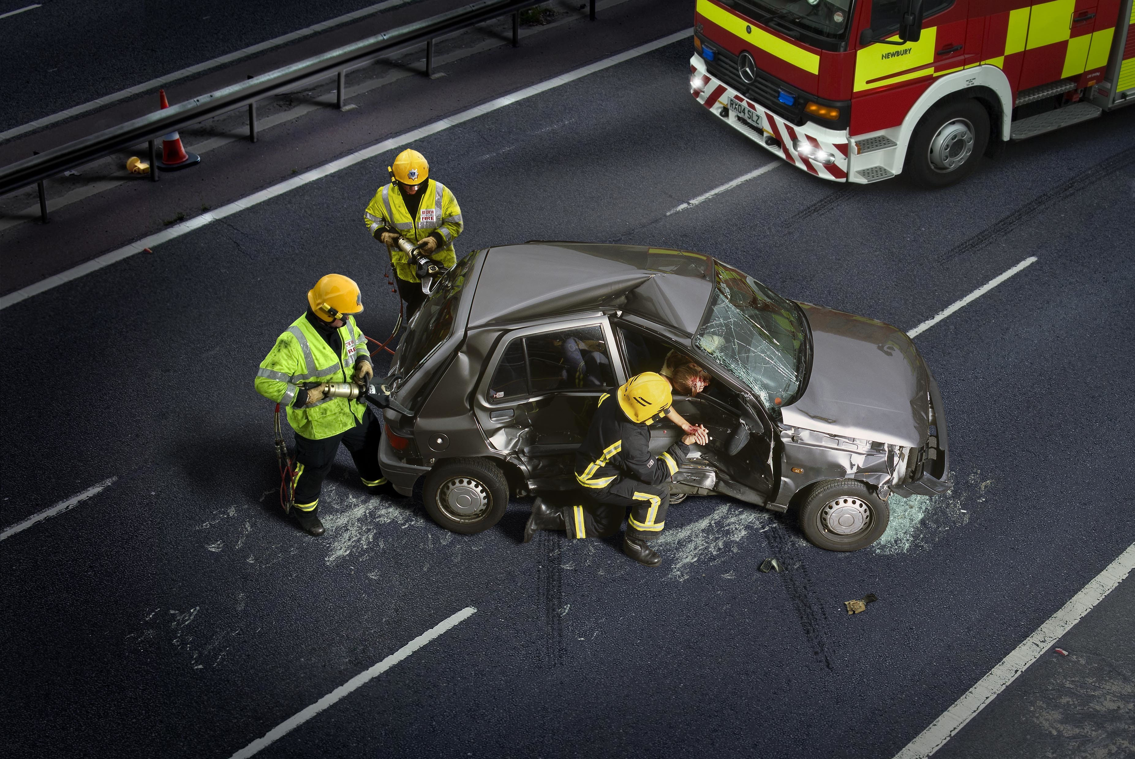 A car smashed up in the middle of a road. Three fire fighters stand by the wreckage.