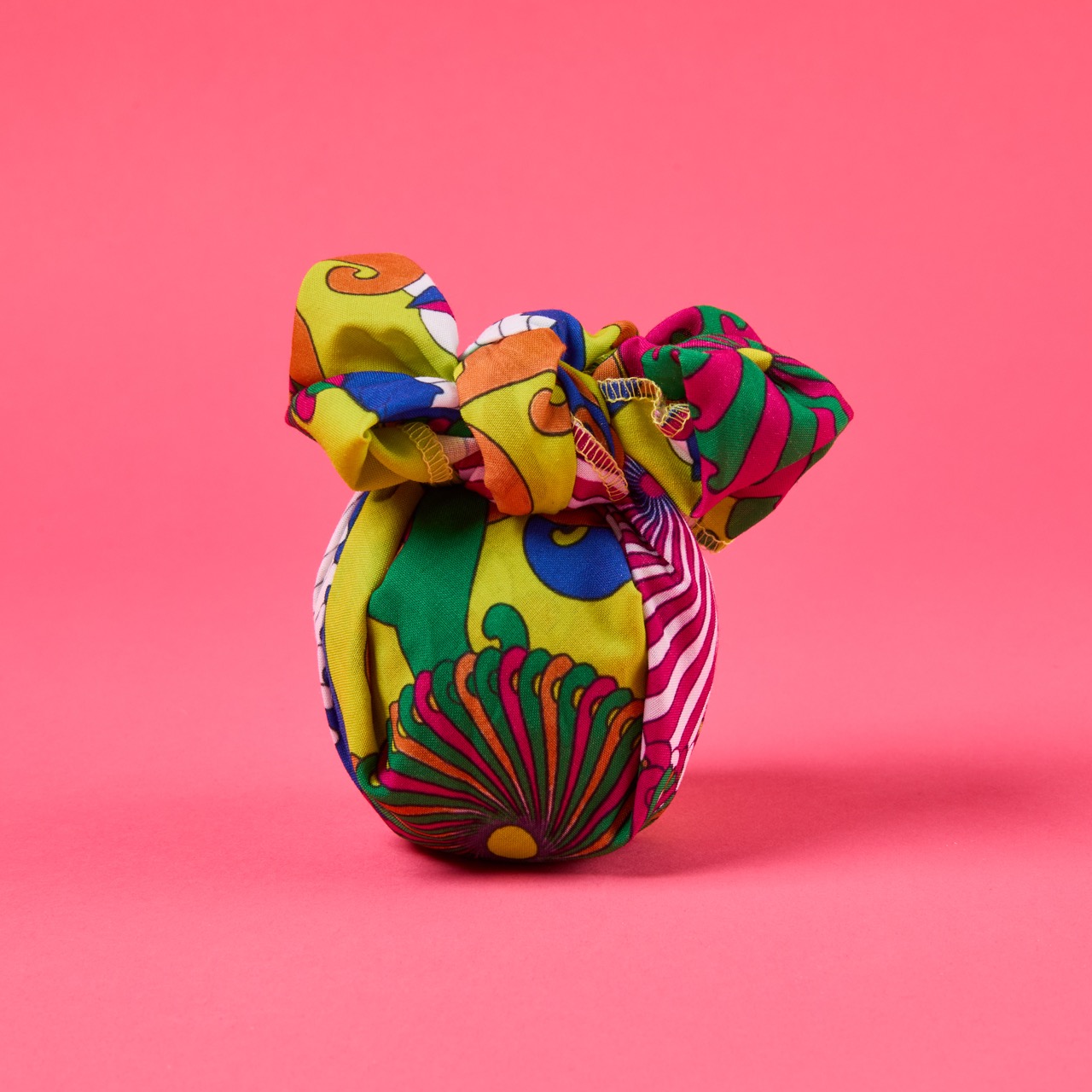Multicoloured, pattered Lush Knot Wrap on a bright coral pink/red background.