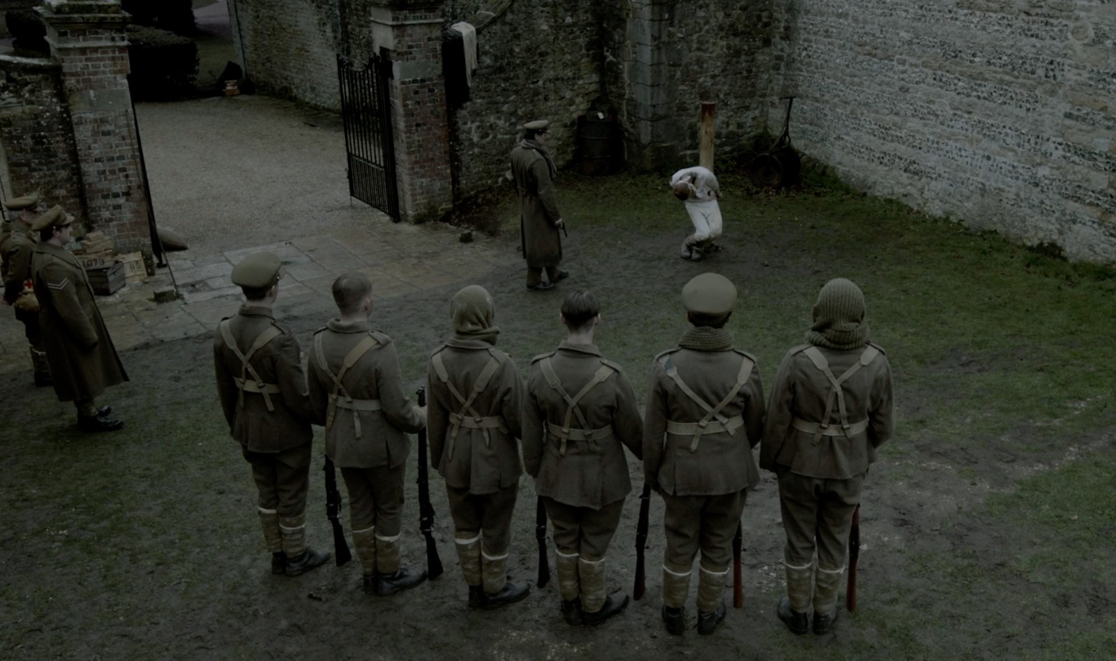 Film still: a group of soldiers lined up in muddy abandoned castle grounds, staring at a tied-up hostage in white.