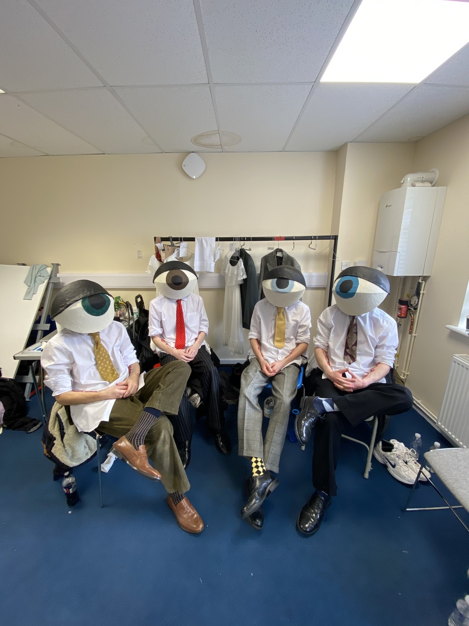 Four actors sitting in shirts, ties and trousers, and wearing headgear resembling eyeballs, sitting behind the scenes on "Water into Wine".