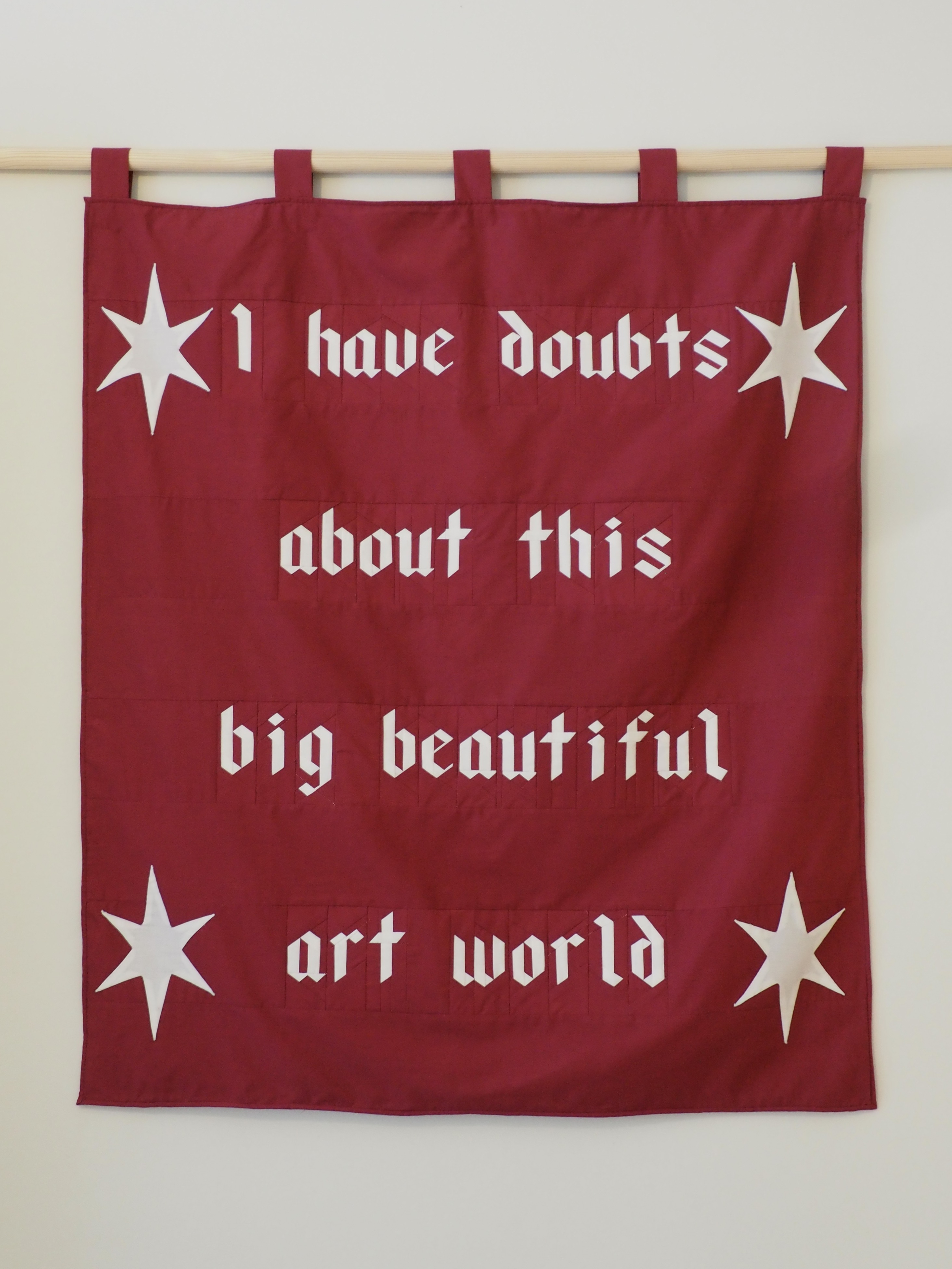 A red piece of fabric hanging on a rod. There are four stars in each corner of the fabric. Gothic text on the fabric reads "I have doubts about this big beautiful art world".