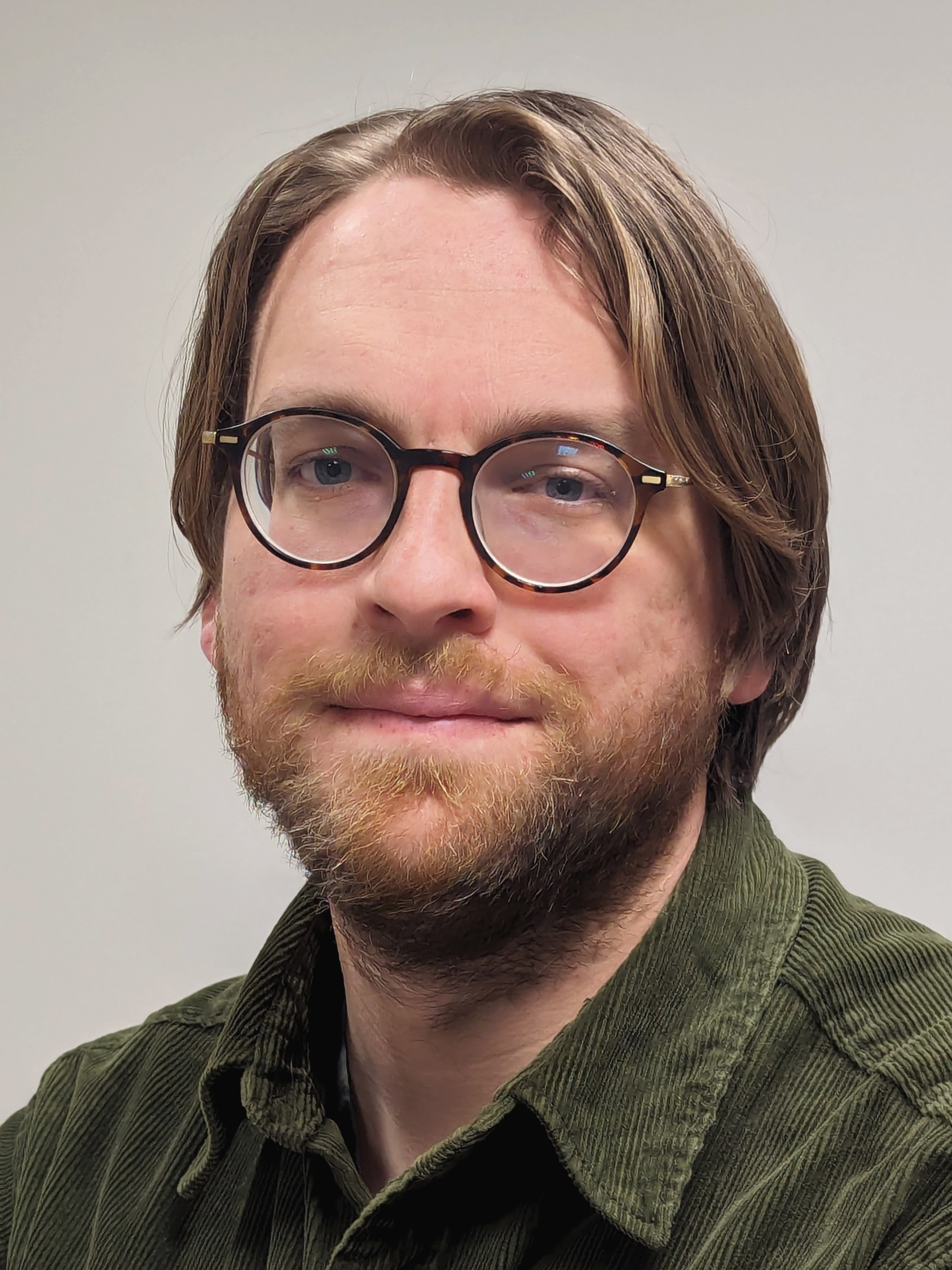 Man with shoulder-length brown hair, round glasses, and beard wearing olive green corduroy shirt against neutral background.