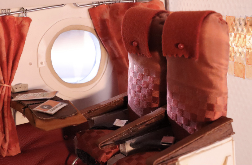 Stop motion model of two seats on an aeroplane with a large round window and a small table.