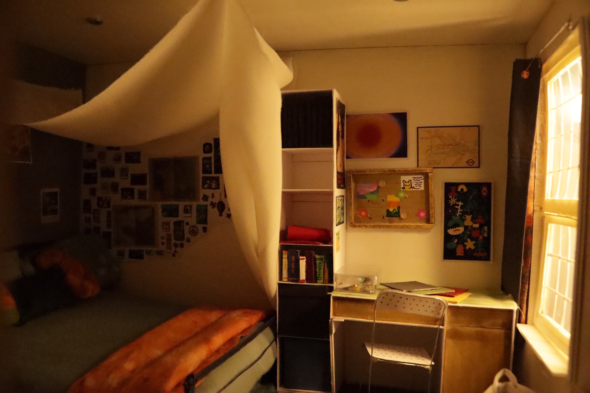 Illustration of a double bed with an orange throw rug at the end, a white canopy above it. Small shelves and a desk are next to the bed. Sunlight shines through the window on the right.