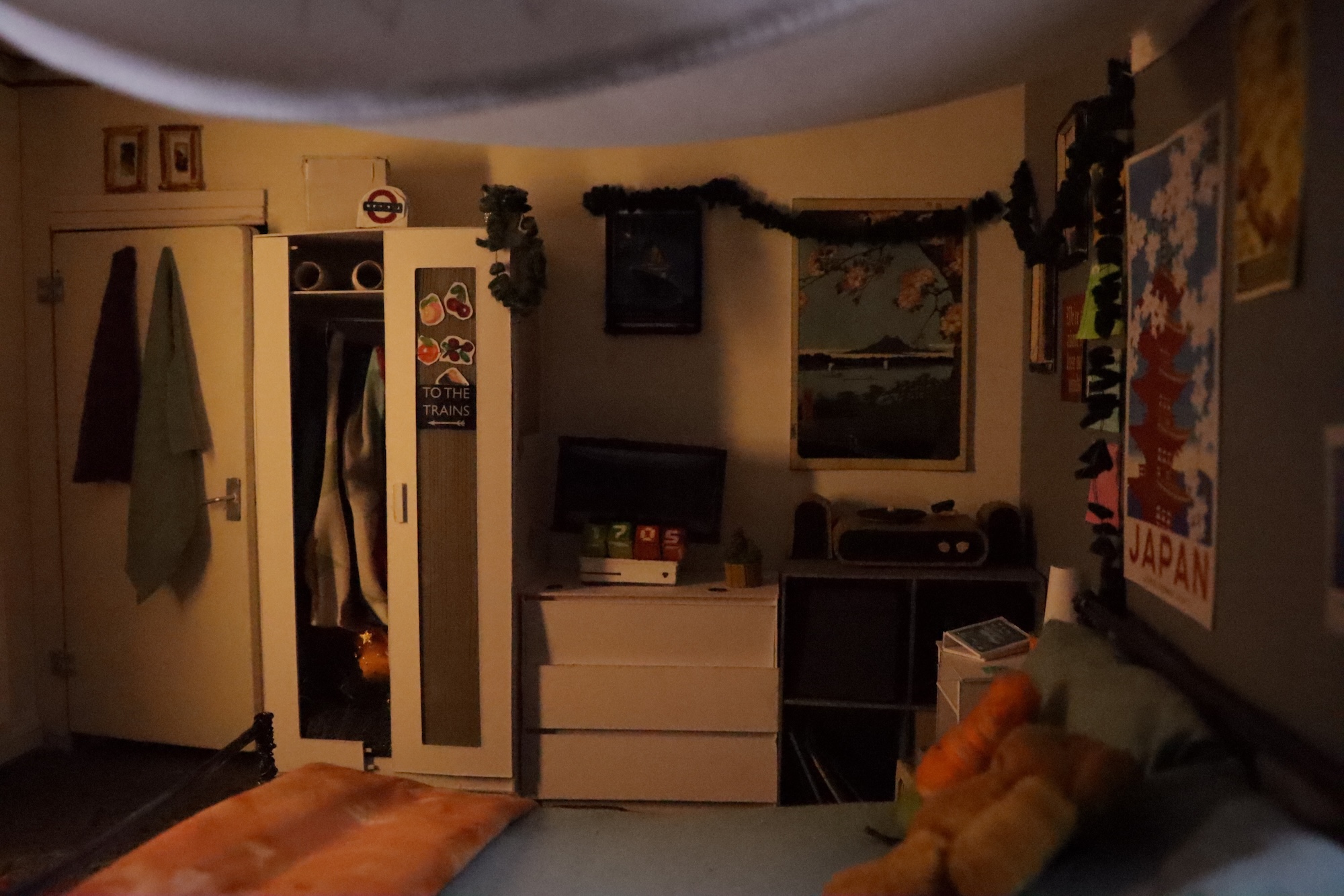 Illustration of a person's bedroom, with a door with towels hung on it, a small wardrobe, a chest of drawers in the background. Posters and artworks hang on the wall. The bed is visible in the foreground.