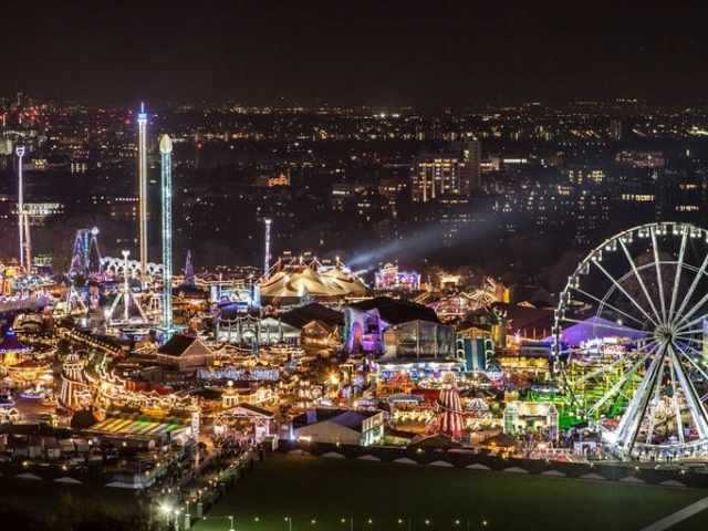 Closer image of a massive bright and colourfully lit fairground viewed at night with a cityscape in the background.