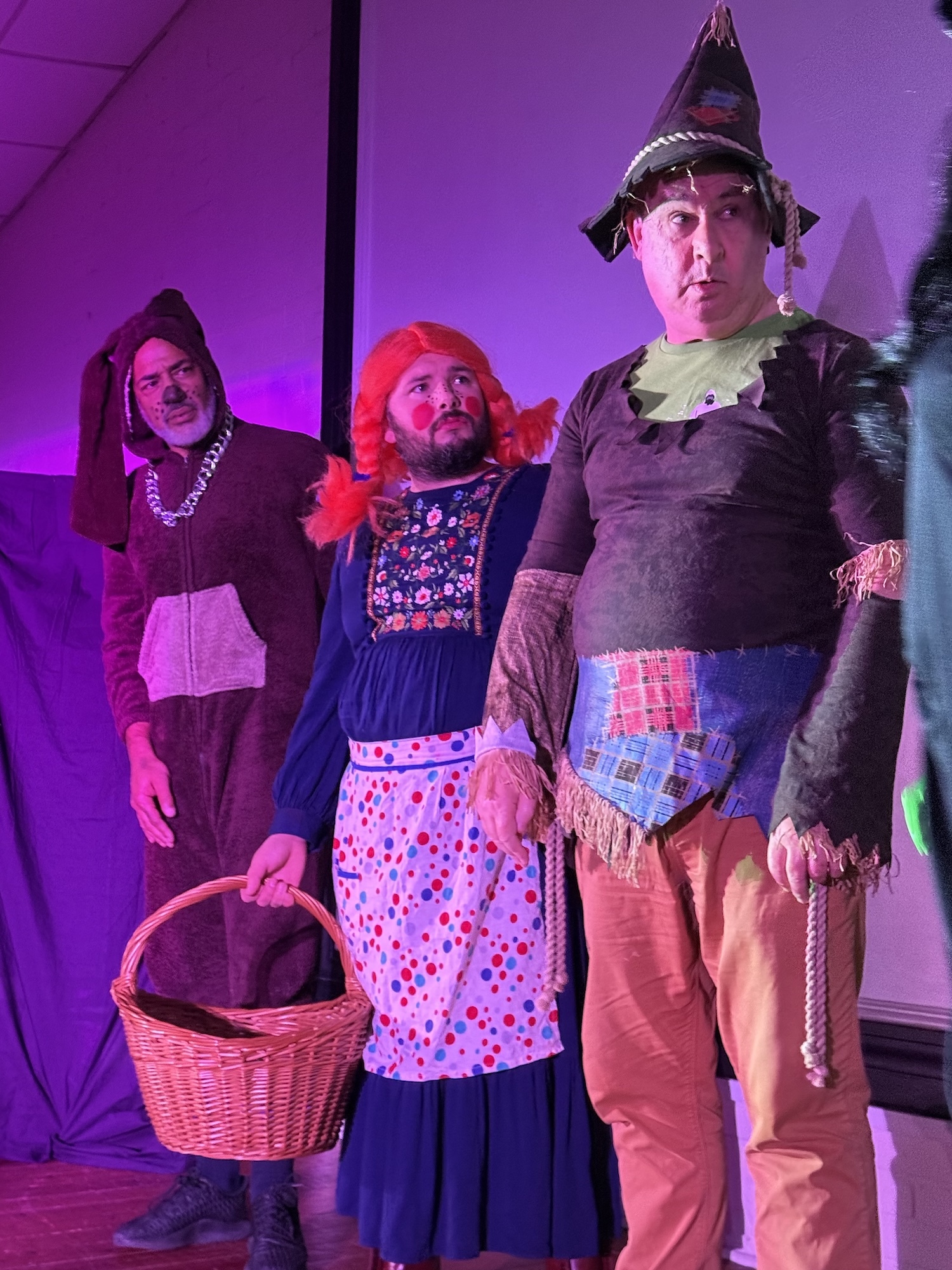 Three actors on stage in costume: woman in black dress with basket, person in blue dress with red headscarf, man in medieval armour with pointed helmet. Purple lighting.