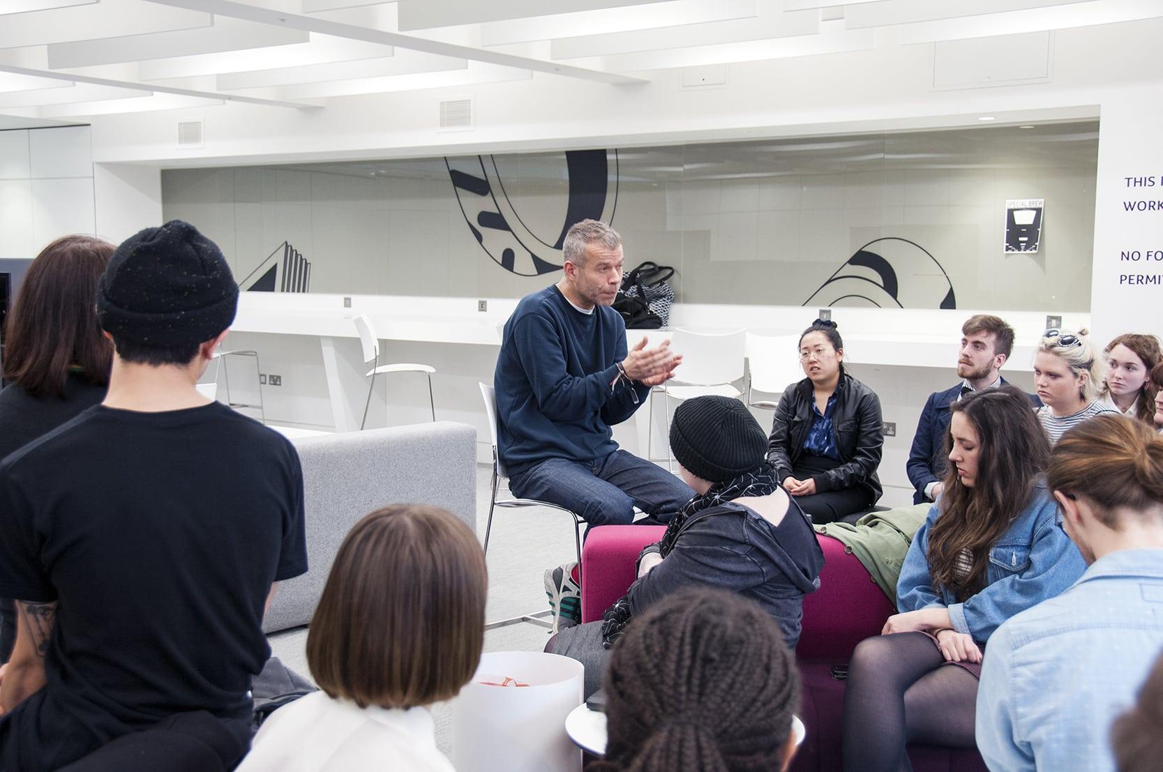 A person sits on a high chair in the centre of a room giving a lecture to a small crowd seated in front of him.