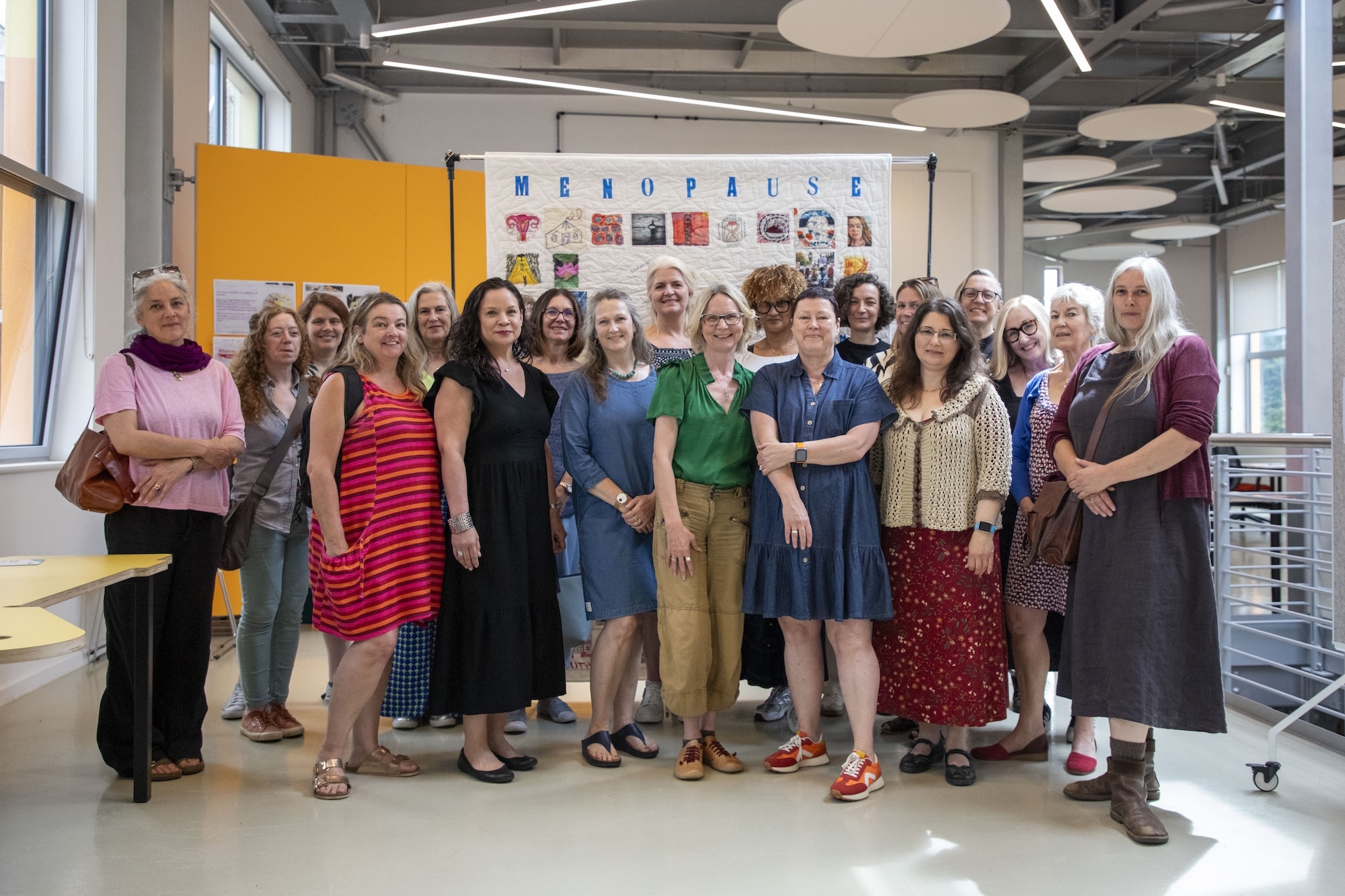 Large group of women standing together in modern office space with exposed ceiling, orange partition wall, and colourful display boards.