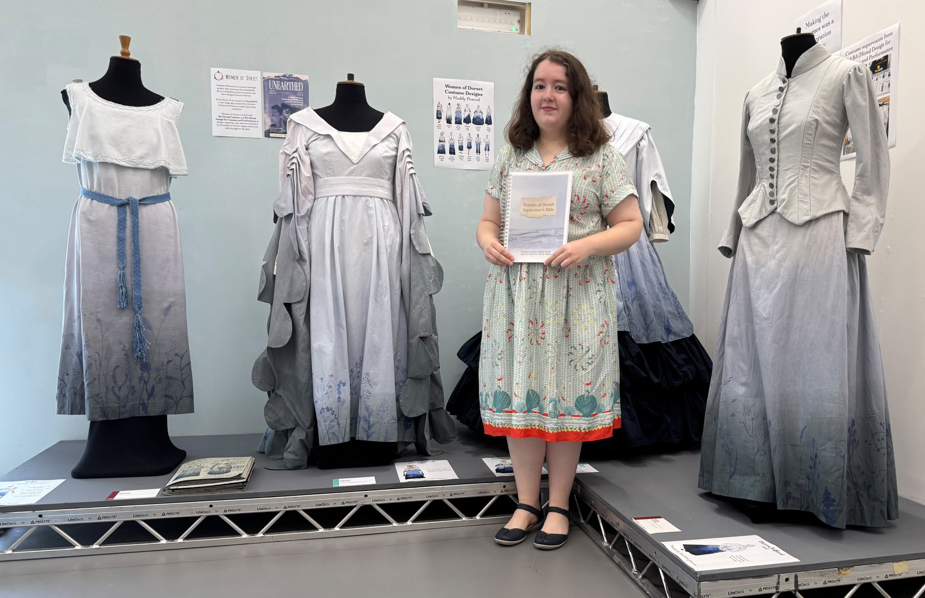 A woman in a floral dress stands holding a document in front of a display of vintage dresses on mannequins. The dresses are in shades of grey and blue, with various designs, showcased on a low platform in a light-coloured room.